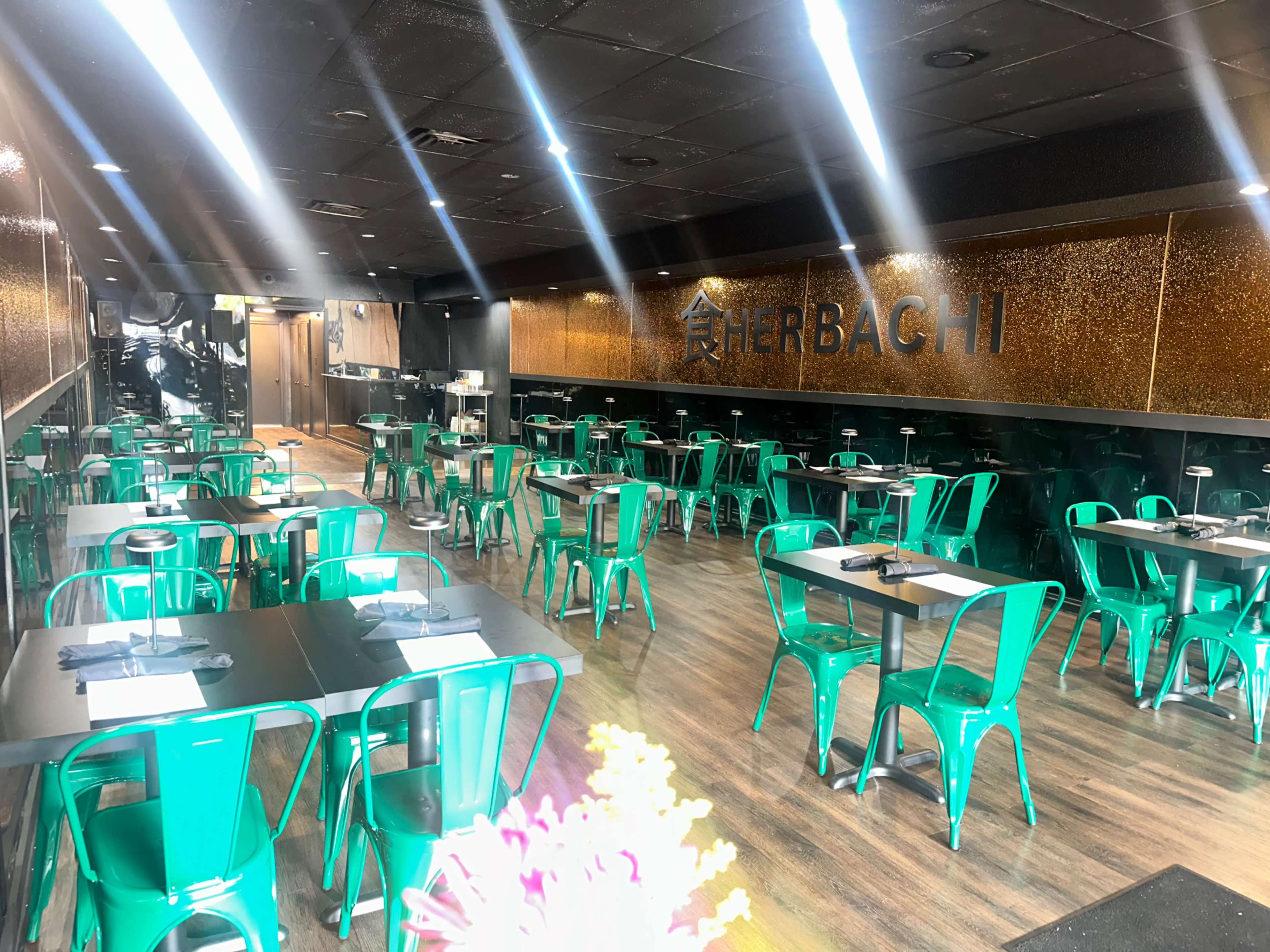 The image shows an empty restaurant interior with green metal chairs and black tables, featuring a large wall sign that reads "HERBACHI."