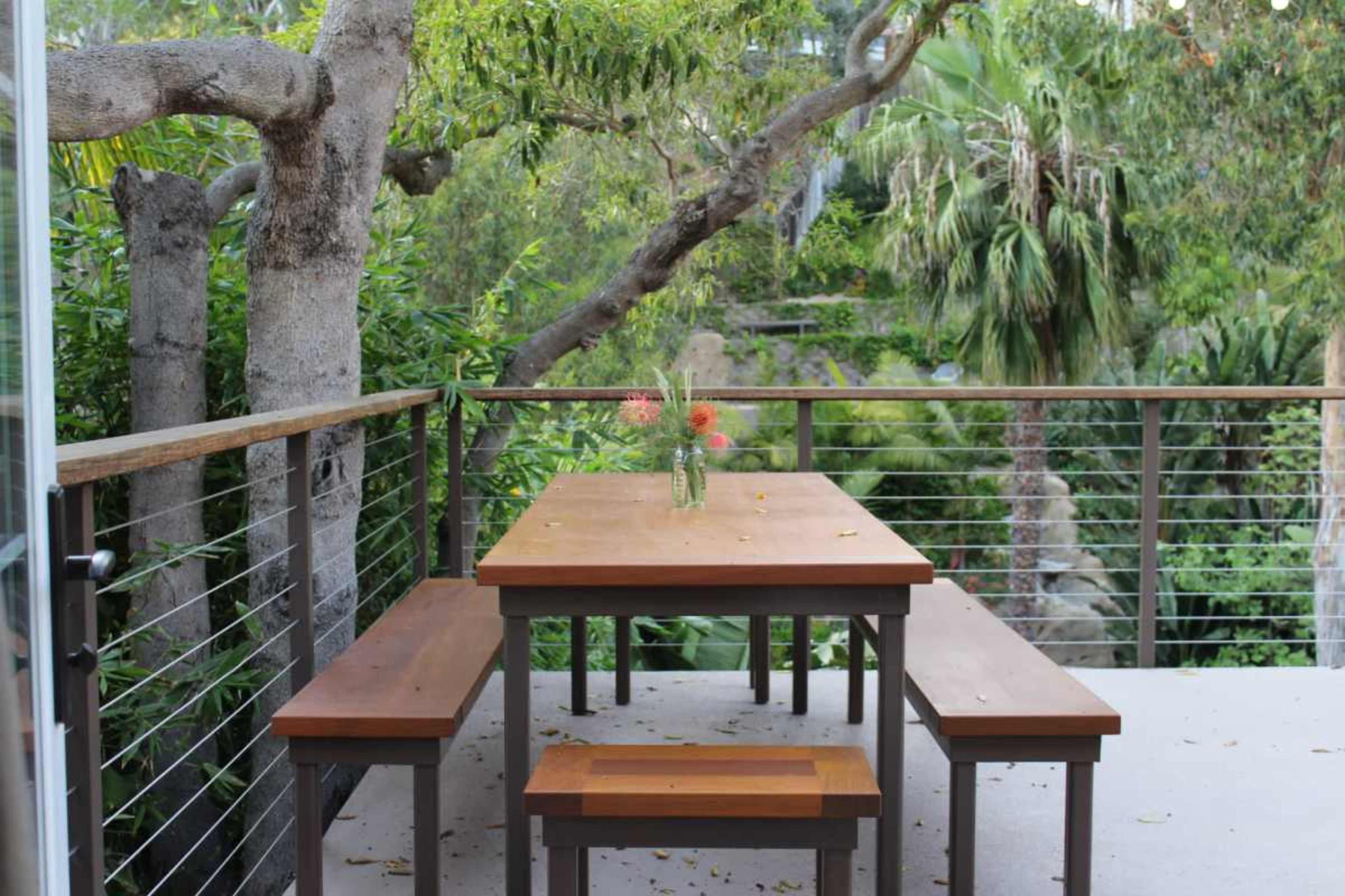 A wooden dining table with benches is set on a patio surrounded by lush greenery and trees.