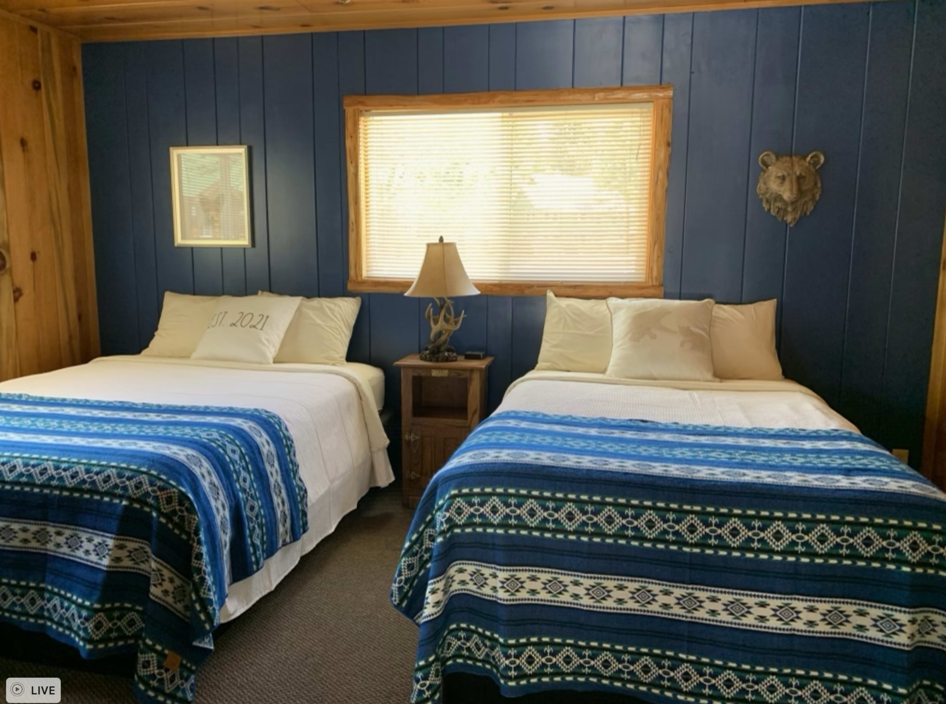 A room with two beds covered in blue and white patterned quilts, a nightstand between them, and a window with blinds.