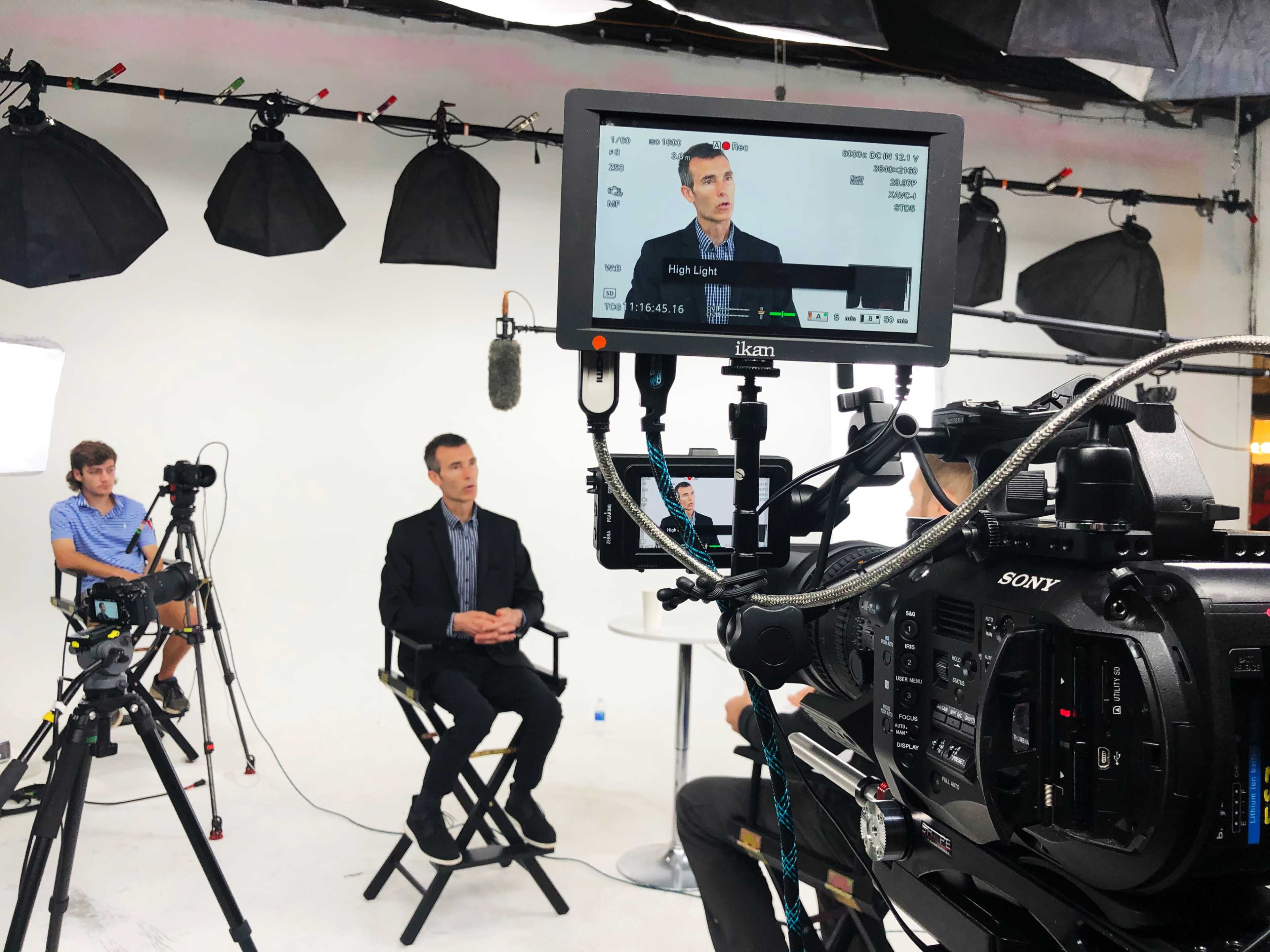 A man in a suit sits on a chair in a well-lit studio, while a person operates a camera and other filming equipment.