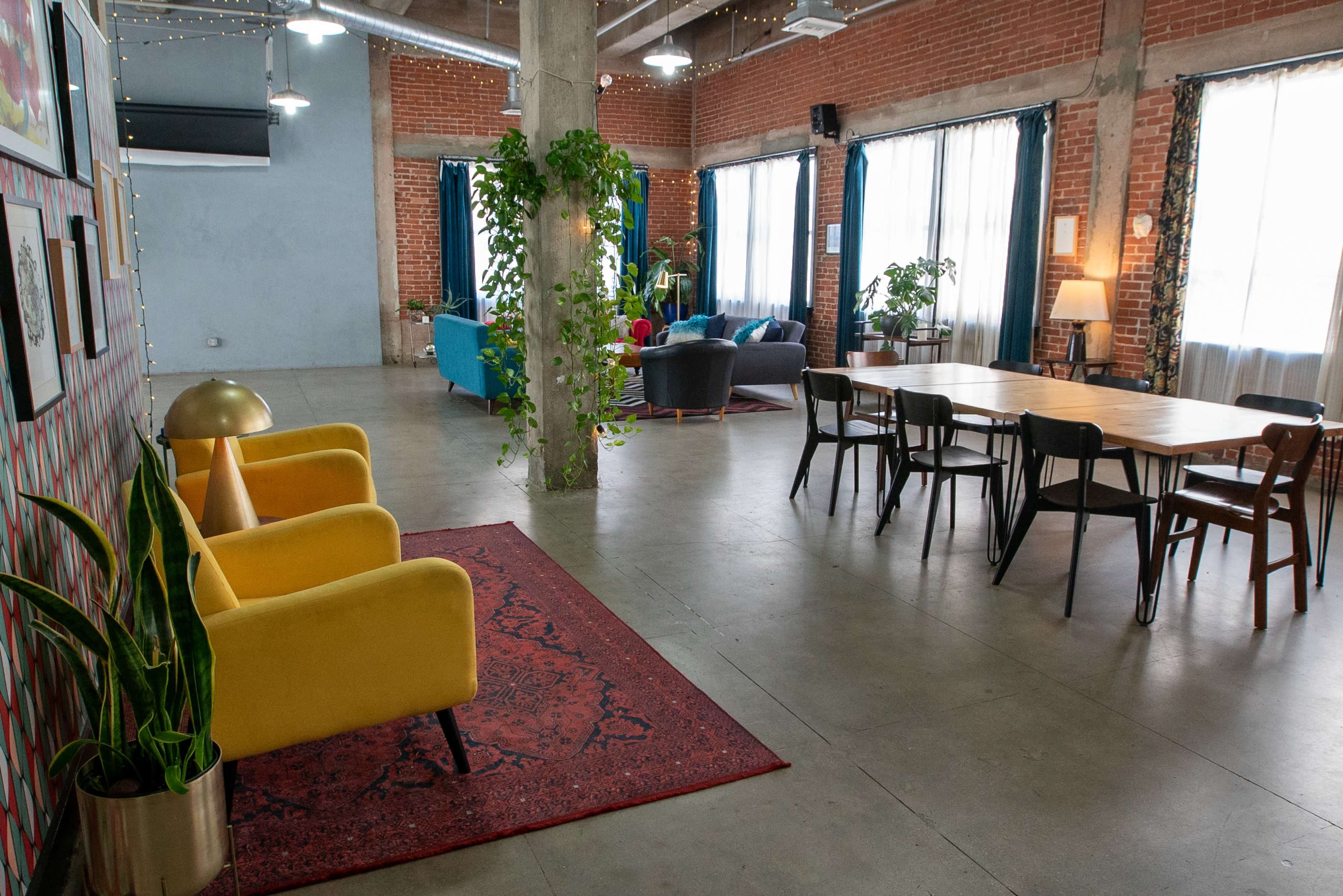 A modern, open space featuring a mix of seating areas, a dining table, and large windows with plants throughout.