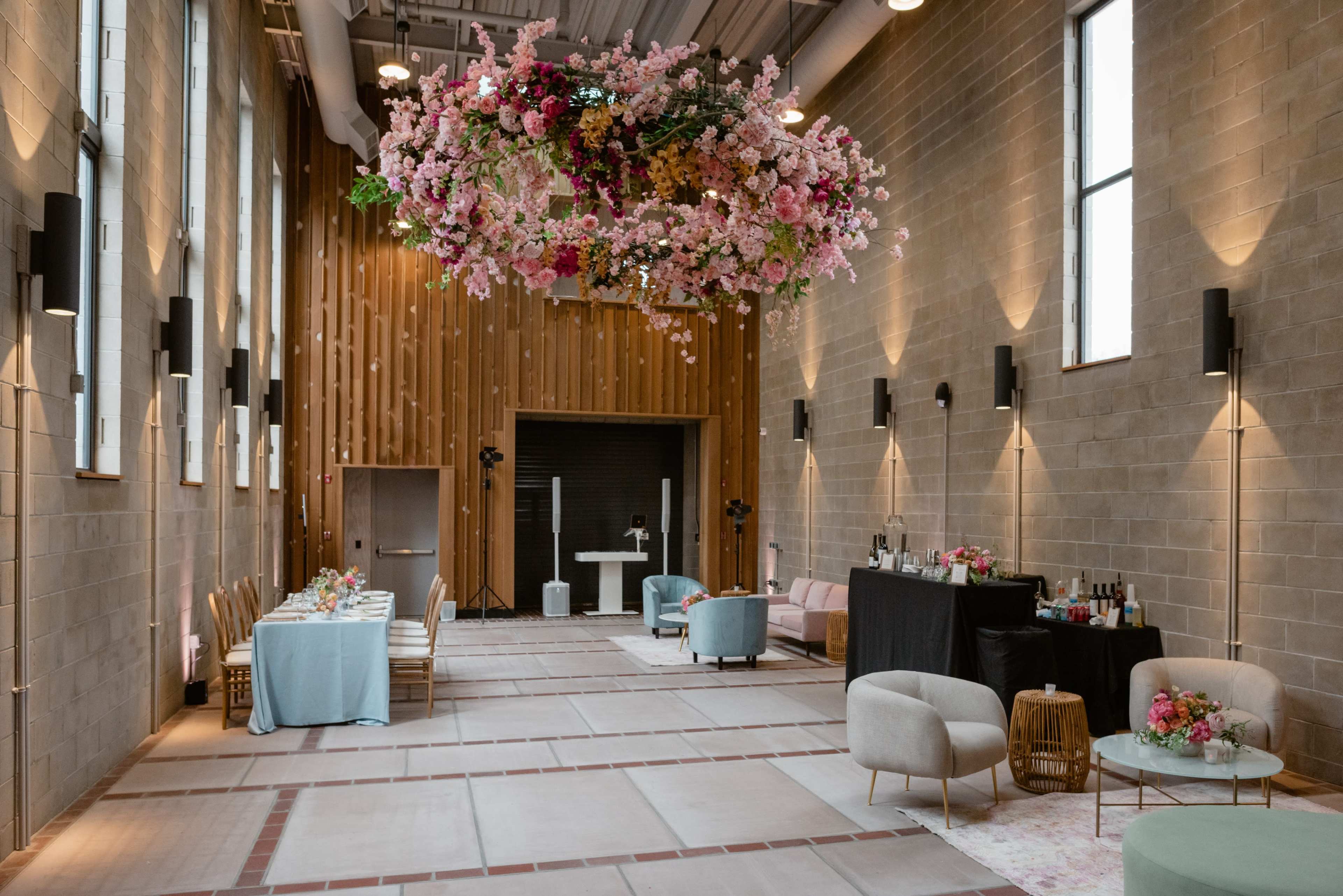 A spacious venue features a long dining table, a bar setup, and a floral arrangement suspended from the ceiling amidst stone walls and large windows.