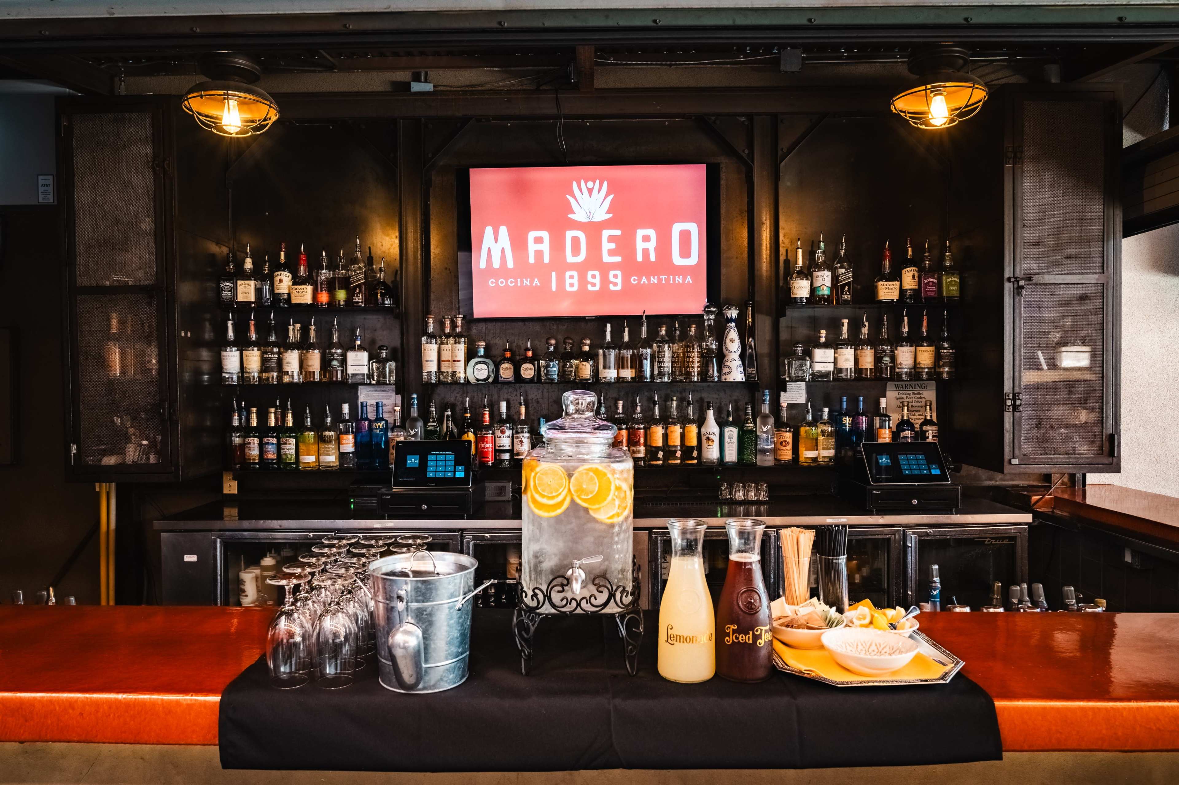 The image shows a bar setup featuring a large selection of bottles on shelves, a decorative water dispenser with citrus slices, and various glassware on a countertop.
