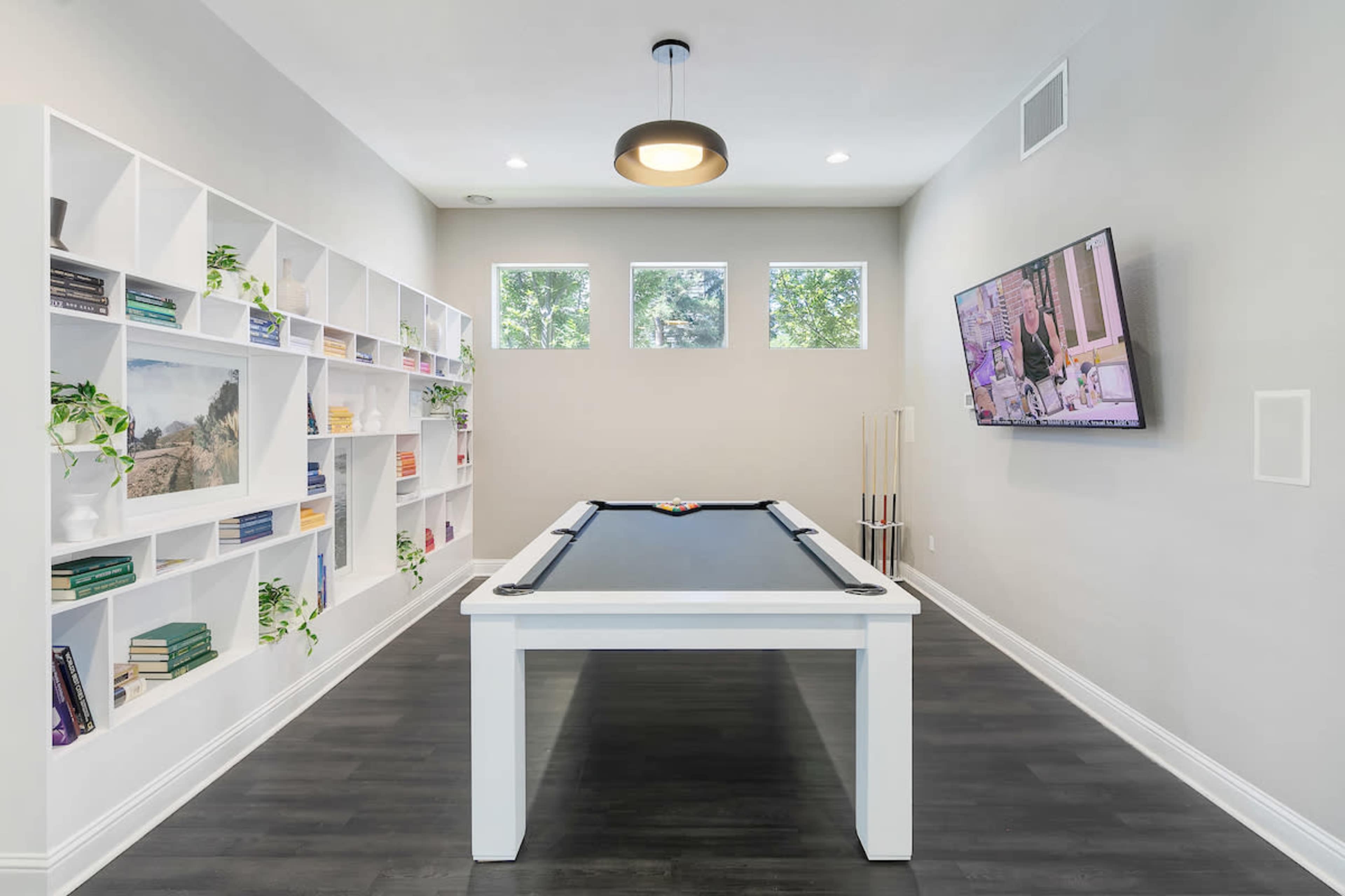 A game room featuring a pool table in the center, surrounded by built-in shelves filled with books and decor, with windows allowing natural light to enter.