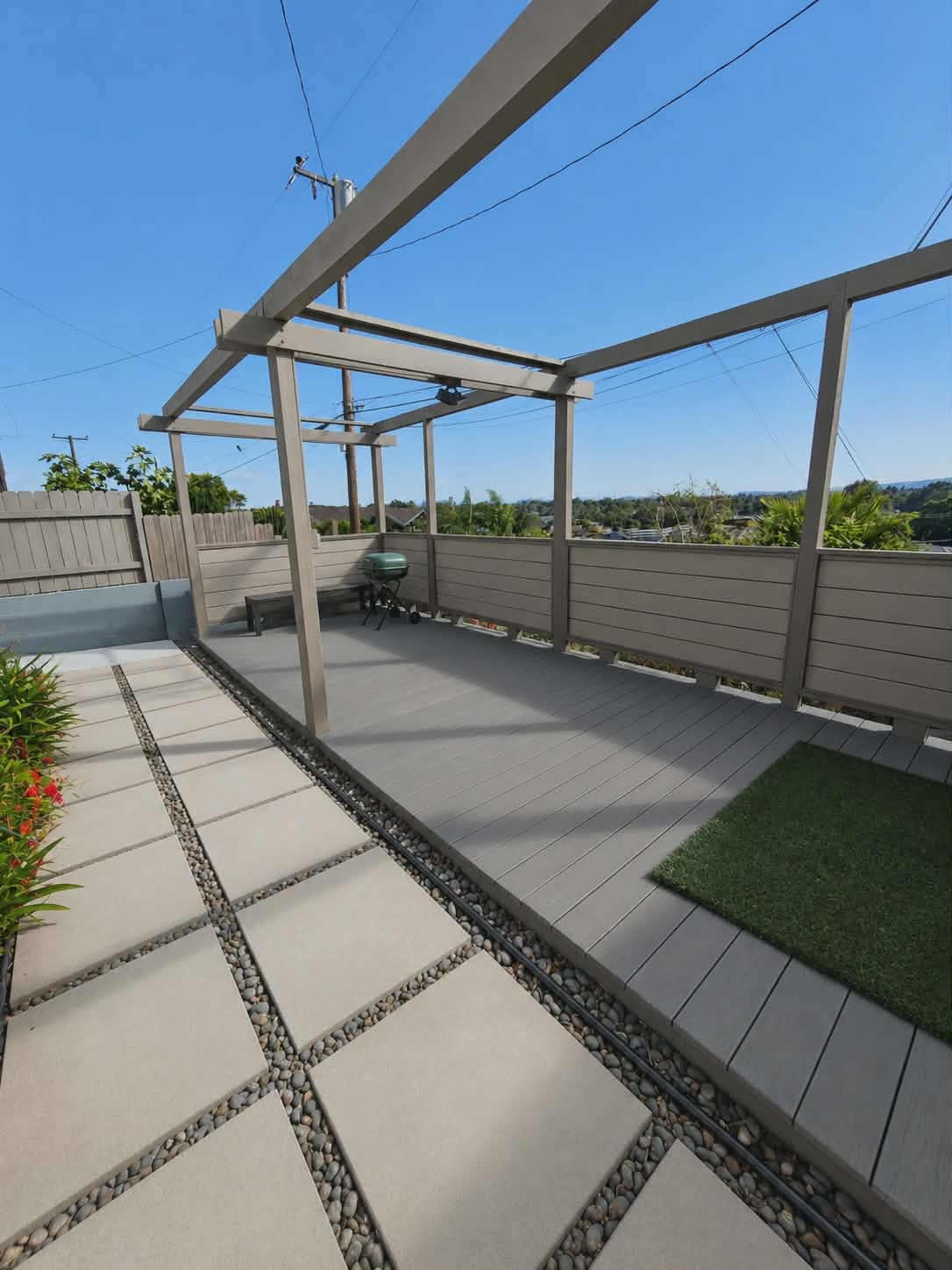 The image shows a modern outdoor patio with a wooden deck, pebble borders, and a clear blue sky in the background.