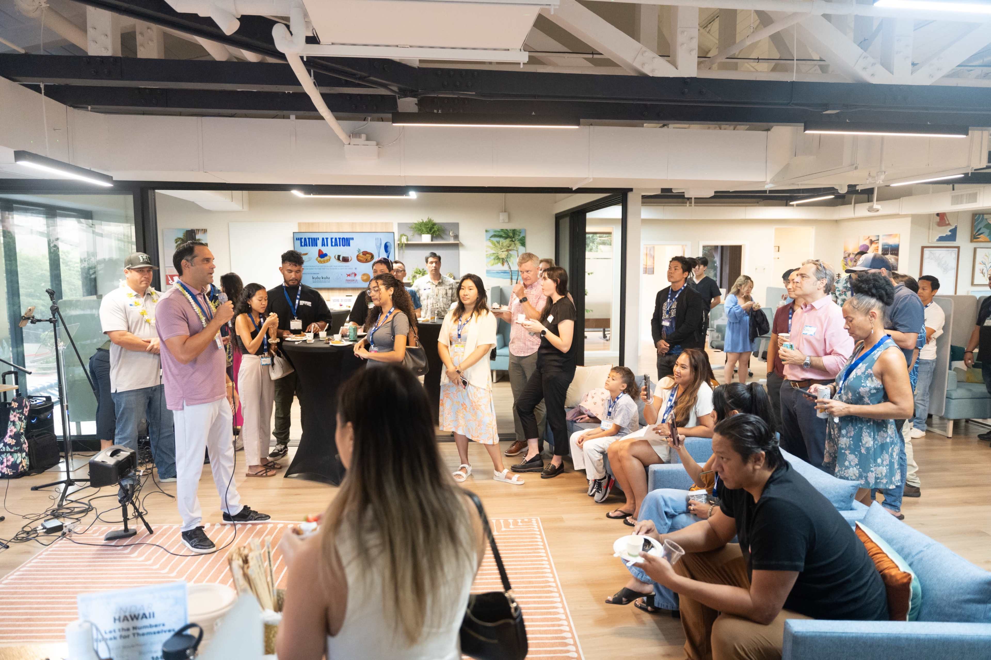A diverse group of people gathers in a modern office space for a community event, with one man speaking at the front near a presentation display.