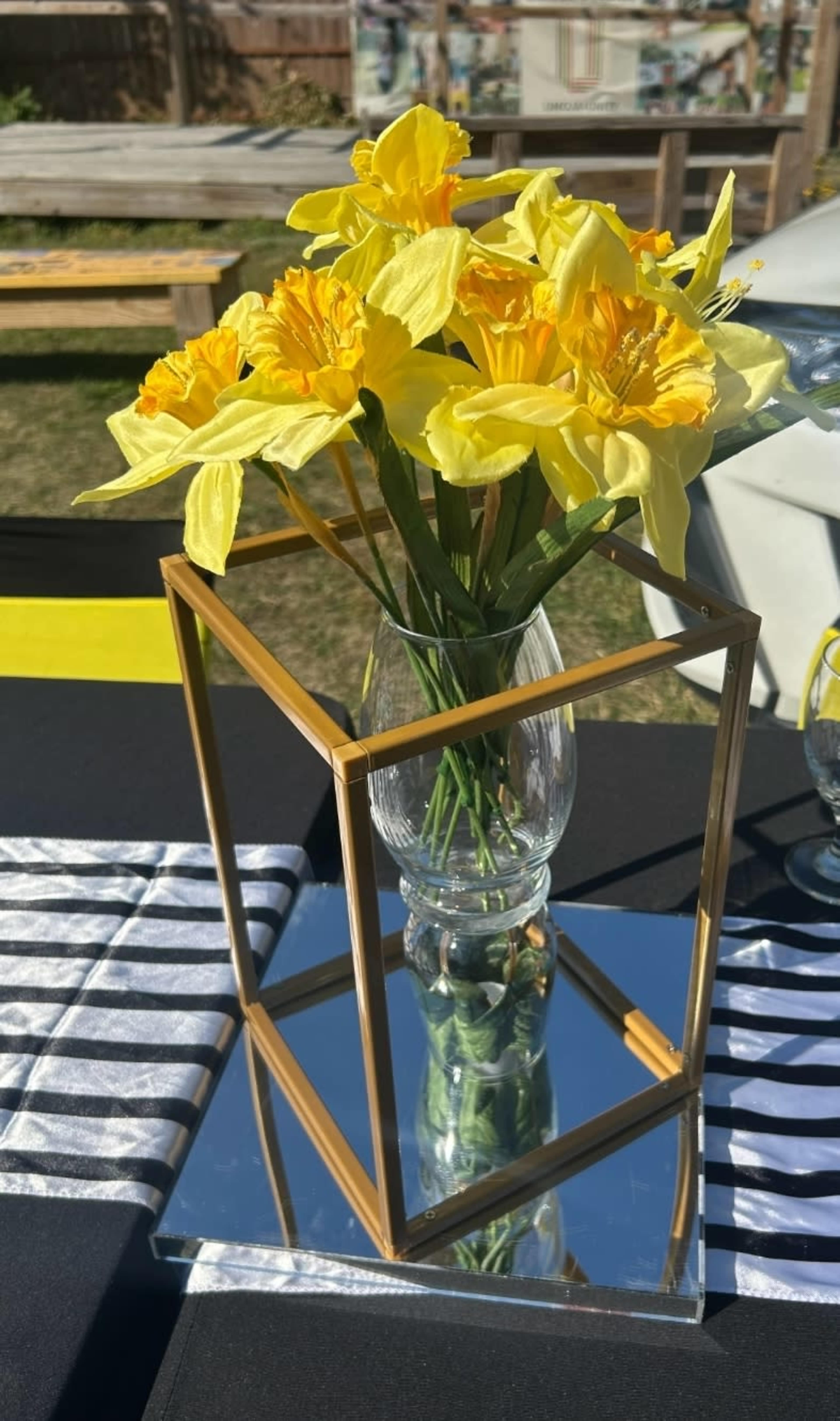 A geometric gold vase holds yellow flowers and rests on a mirrored base atop a striped tablecloth.