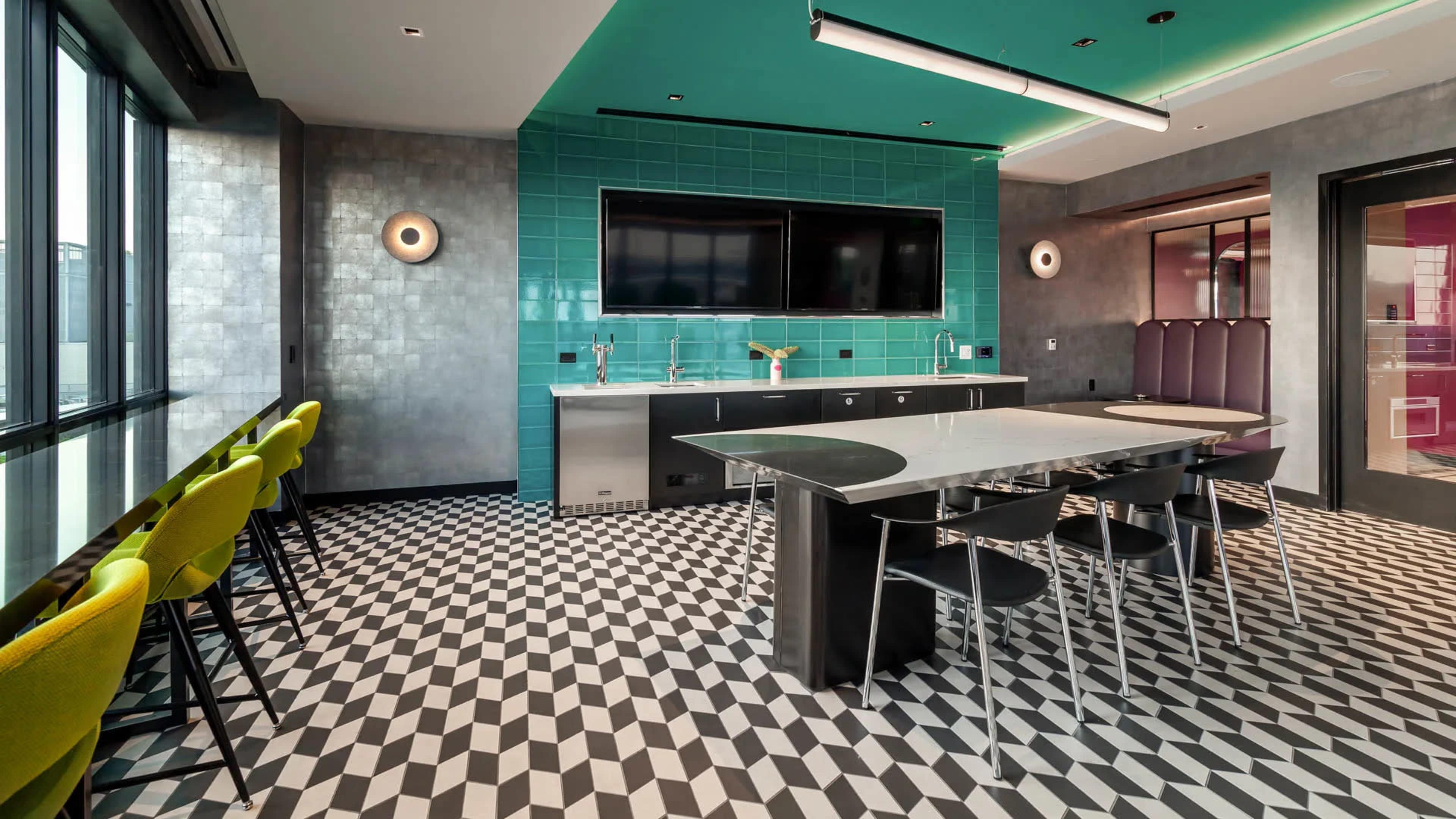The image shows a modern kitchen with teal tiled walls, a large black and white checkerboard floor, a stainless steel countertop, and a seating area with bright green chairs.
