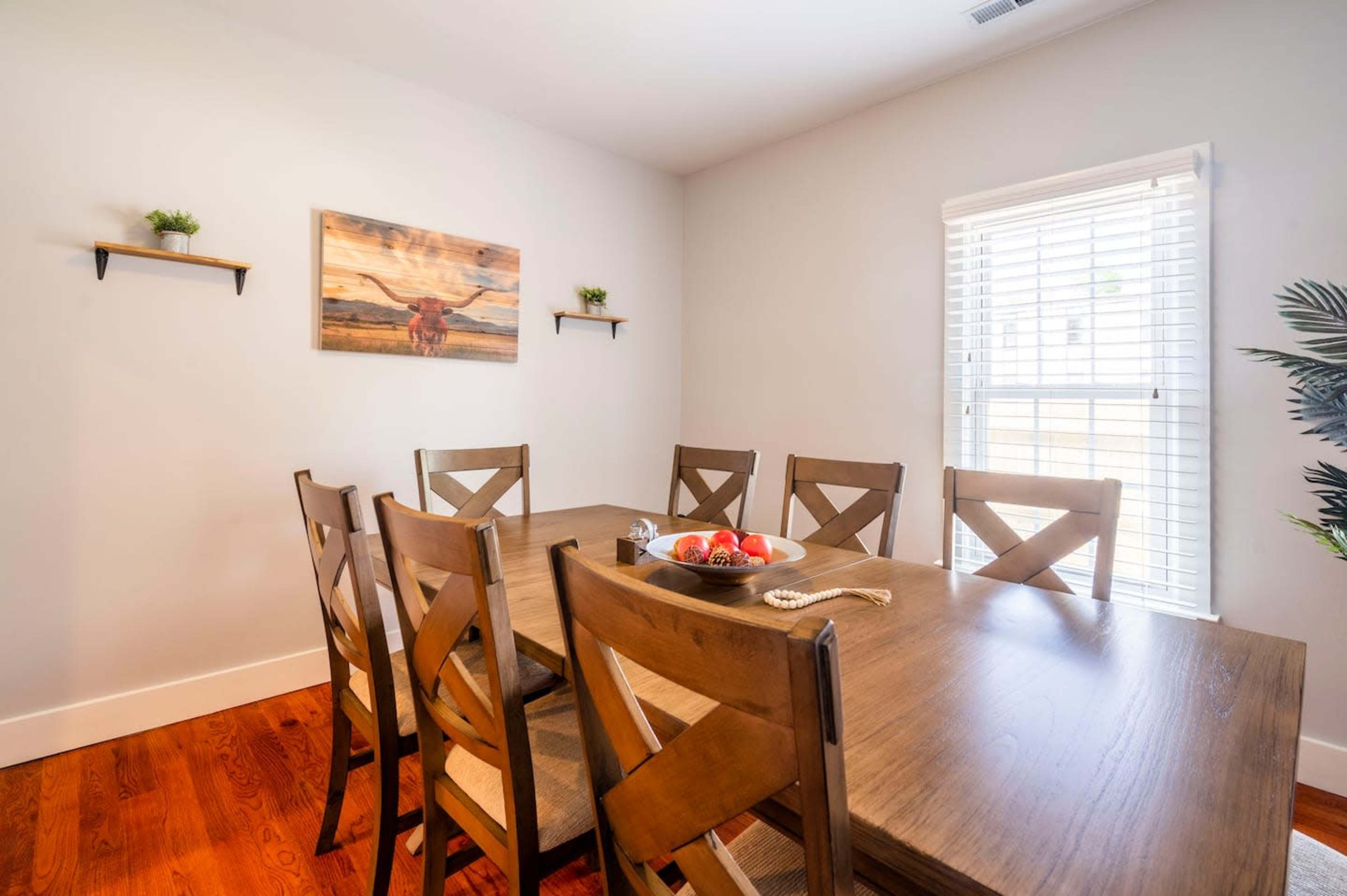 A dining room features a wooden table surrounded by six chairs, with a painting of a horse on the wall and a window letting in natural light.