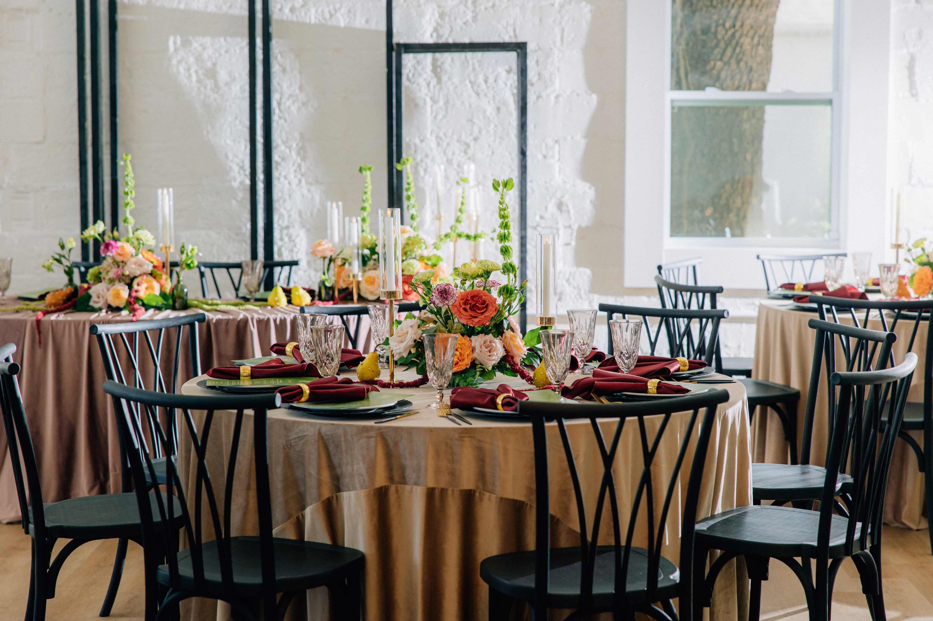The image shows a banquet setup with round tables dressed in different colored tablecloths, each adorned with floral centerpieces and place settings.