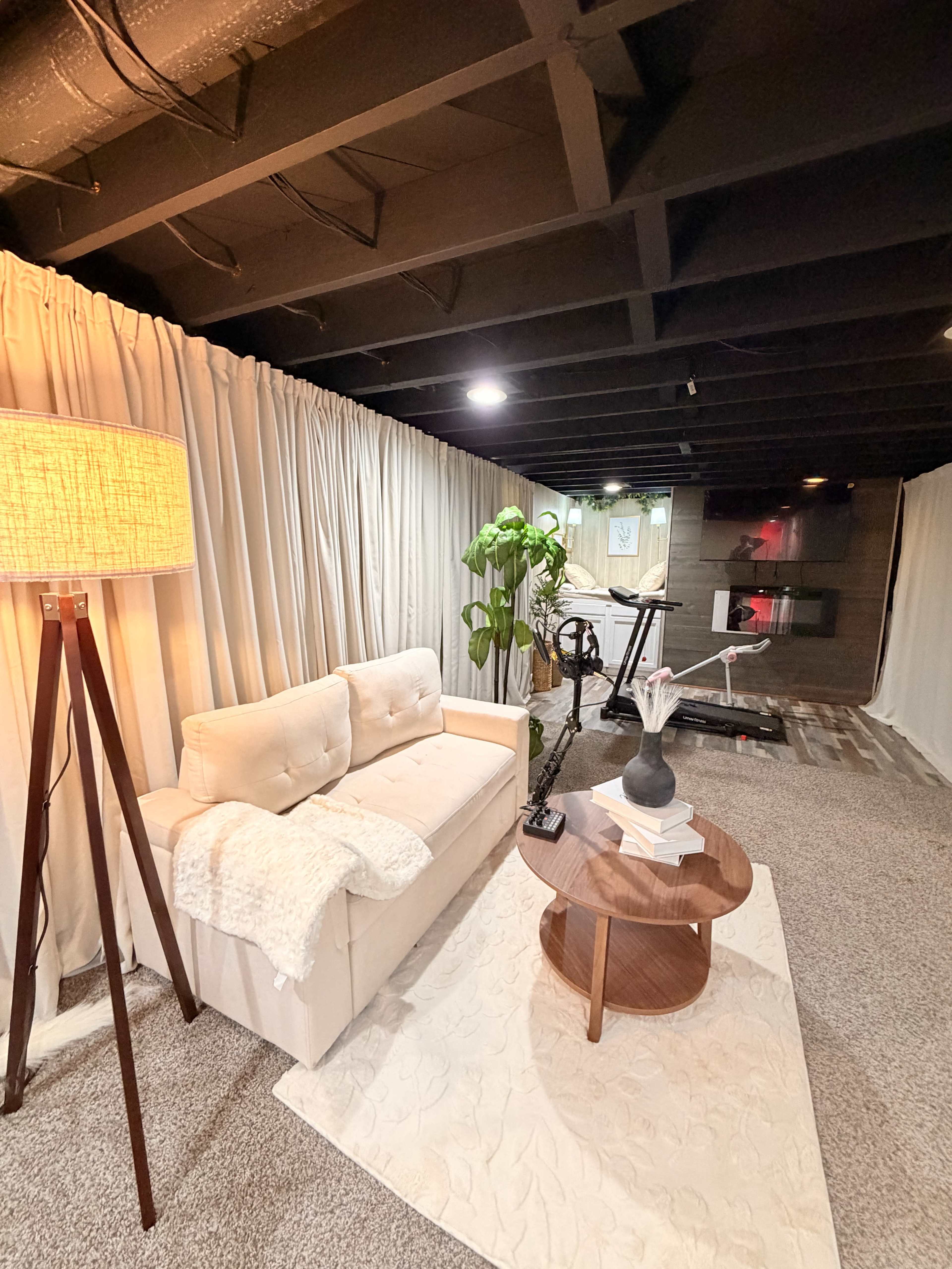 The image shows a cozy basement living area featuring a light-colored sofa, a standing lamp, a small wooden table, and decorative plants against a backdrop of curtained walls.