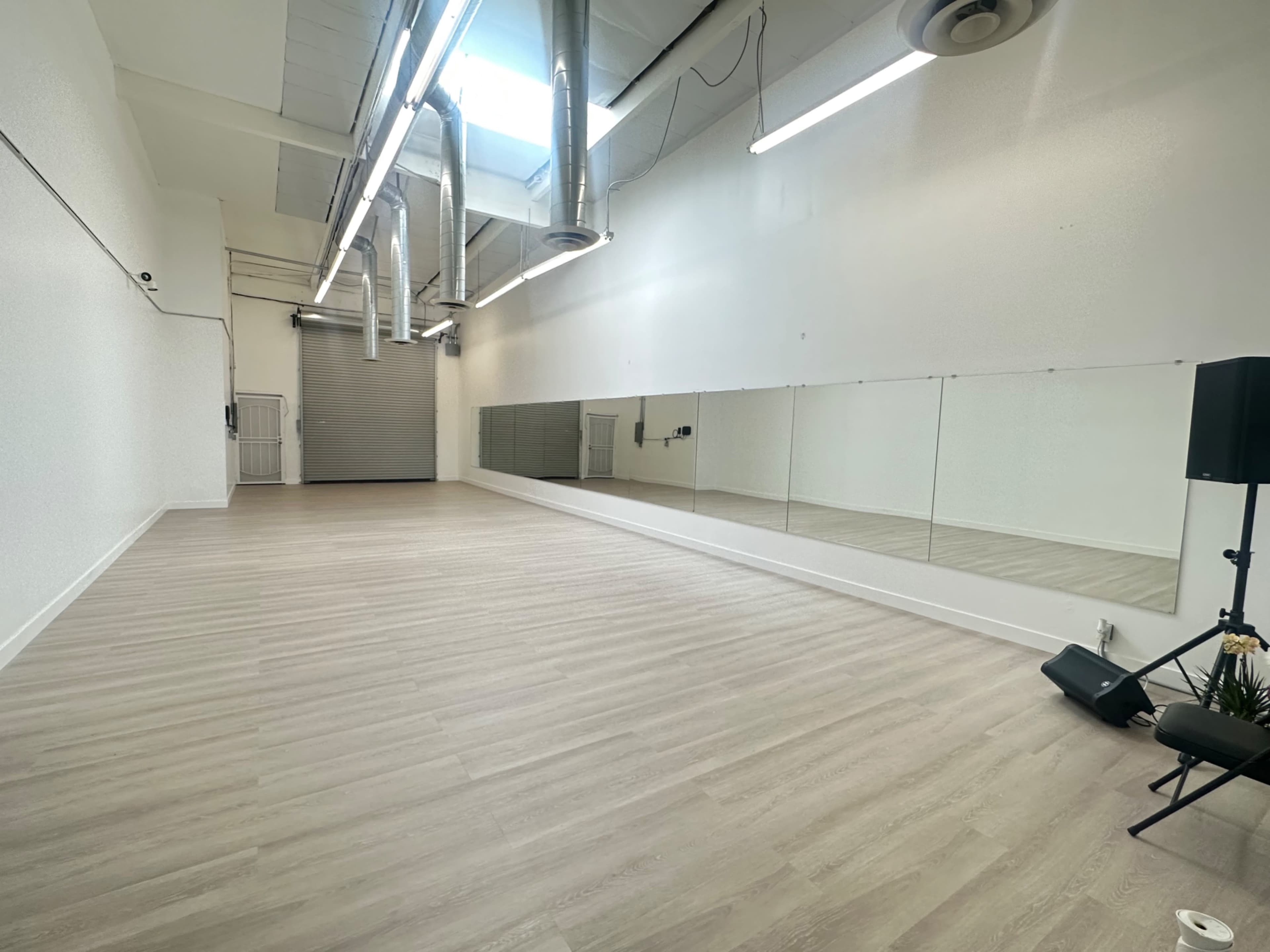 The image shows a spacious, empty studio with light wood flooring, large mirrors on one wall, and industrial-style ventilation ducts overhead.