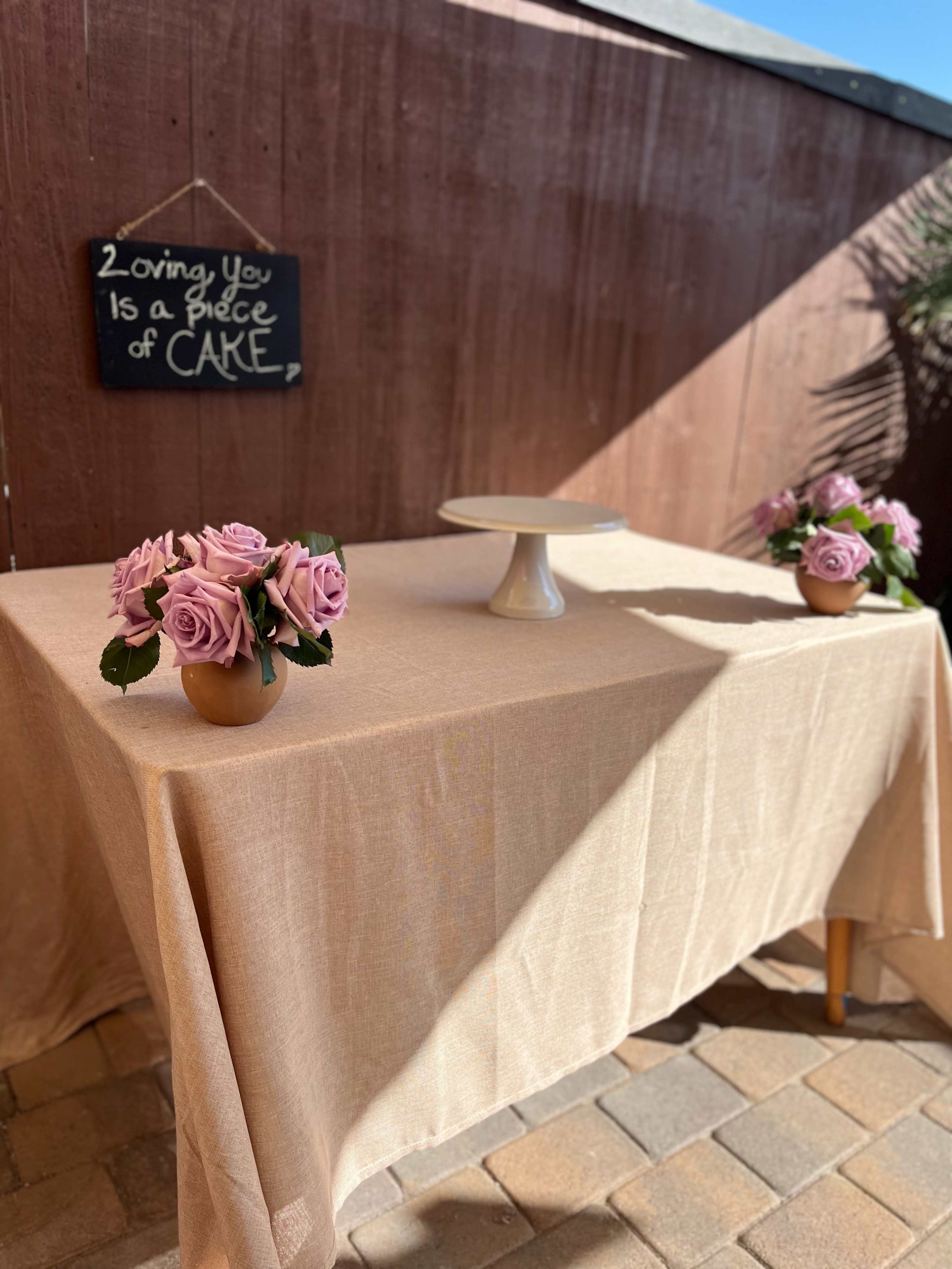 A table with a beige tablecloth is set outdoors, adorned with pink roses in vases and a cake stand, alongside a chalkboard sign that reads "Loving You Is a Piece of Care."