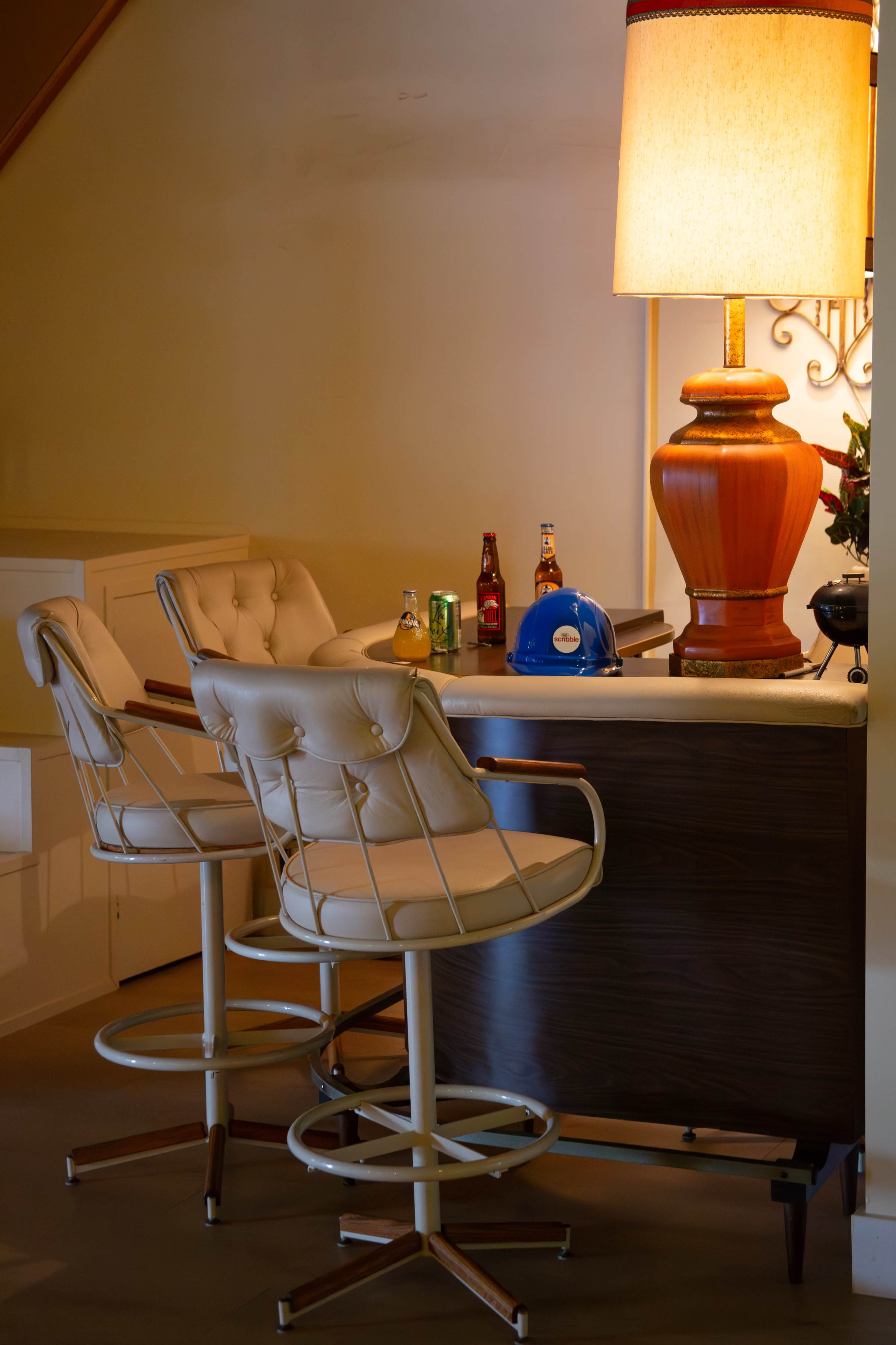 A small bar area with two white swivel stools, a large lamp, and several bottles of beverages on the counter.