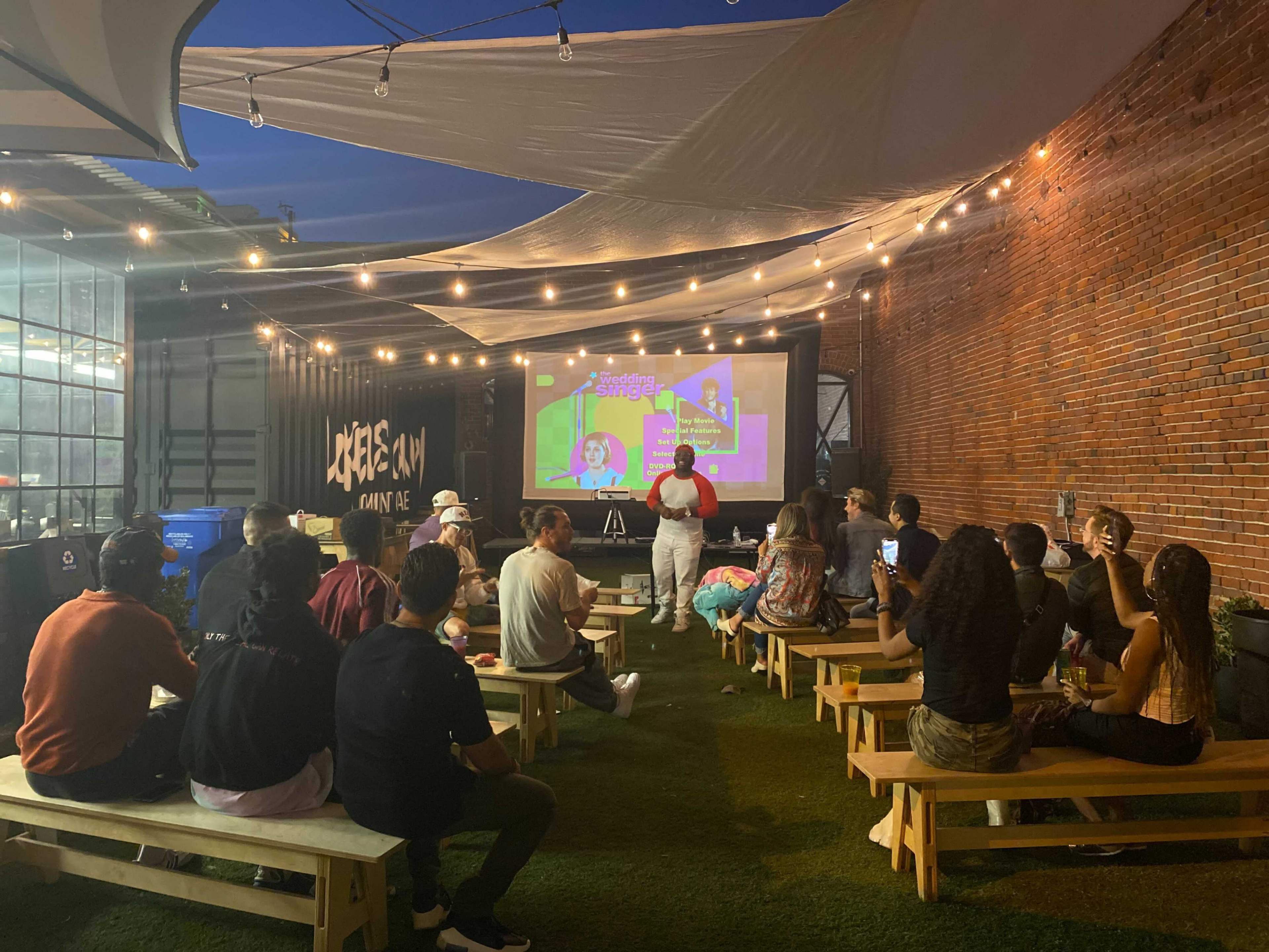 A group of people are seated on benches in an outdoor space, watching a presentation on a large screen illuminated by string lights.