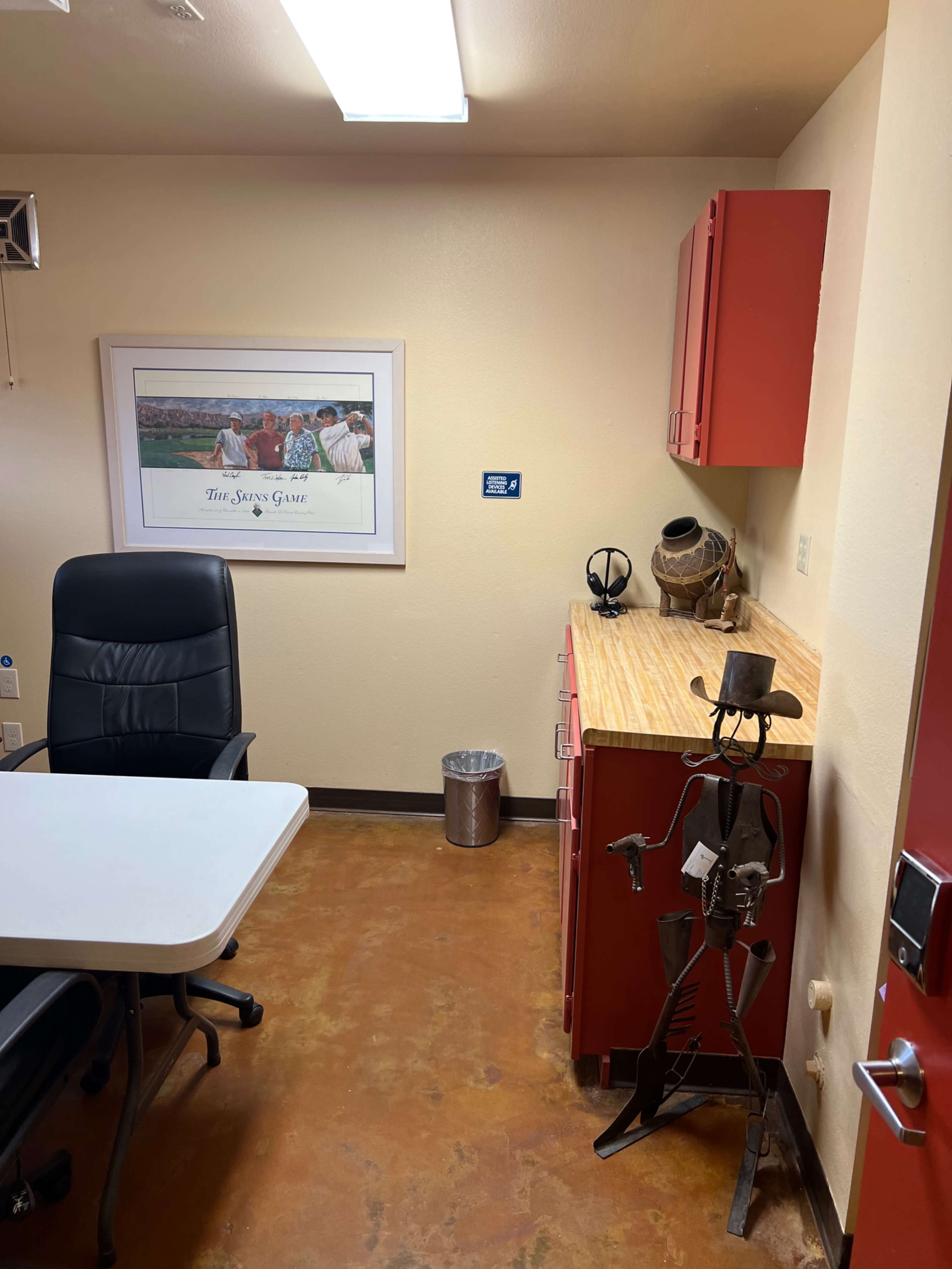 The image shows a small office space with a black chair, a white table, red cabinets, a wooden countertop, a trash can, and a metal sculpture.