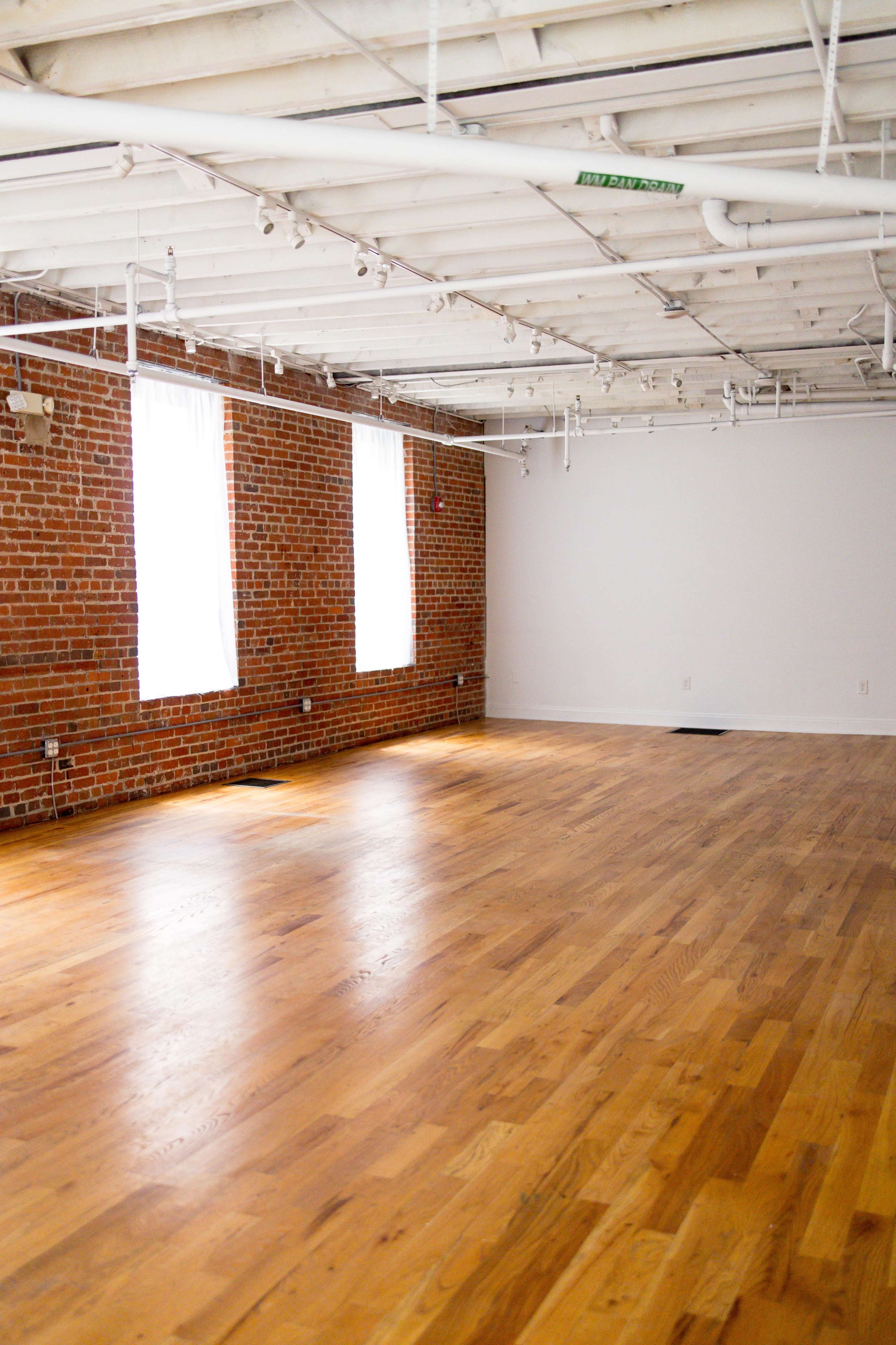 An empty studio space with exposed brick walls, large windows, and a wooden floor.