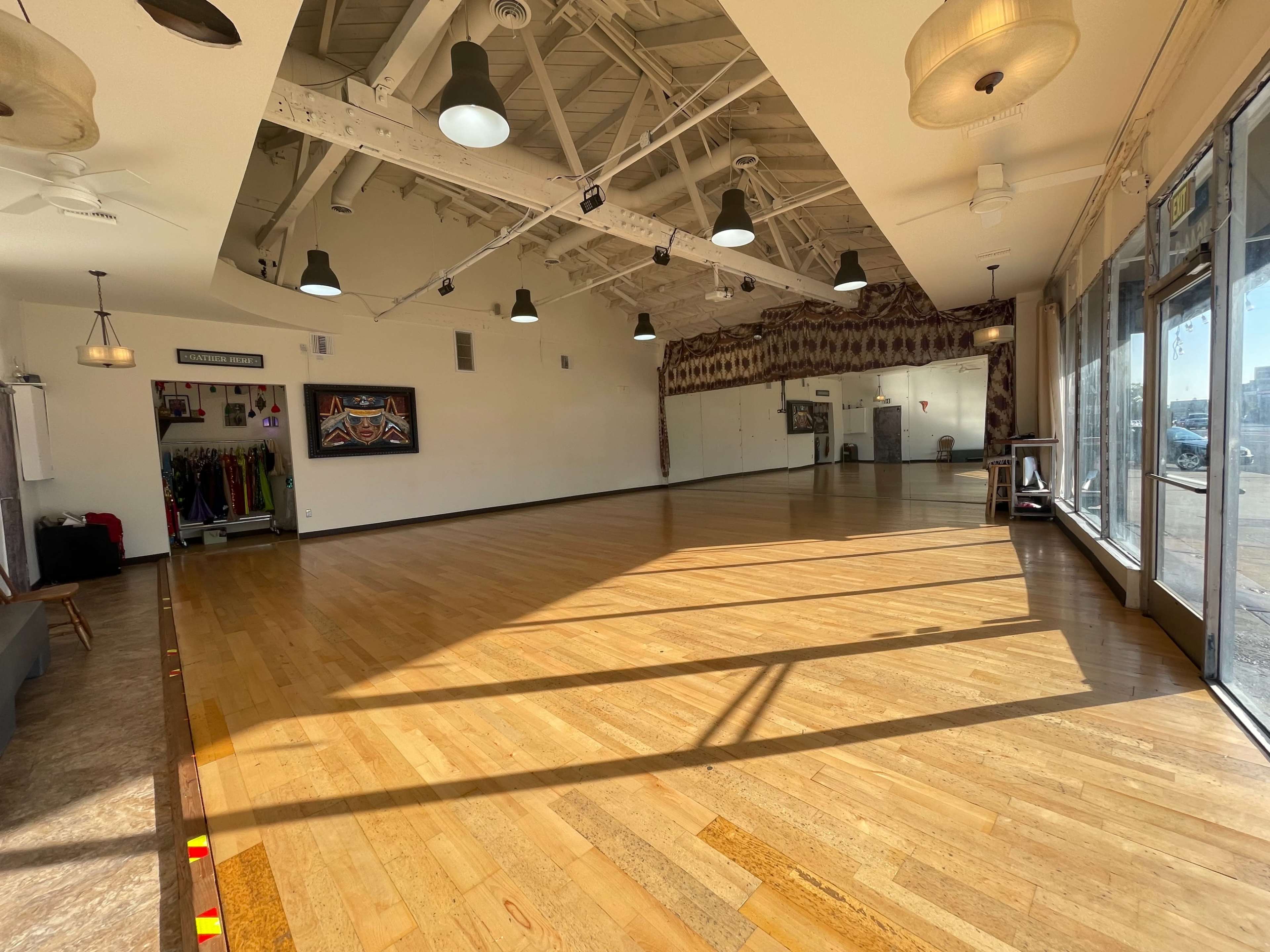 The image shows a spacious, well-lit interior of a dance studio with wooden flooring and large windows.
