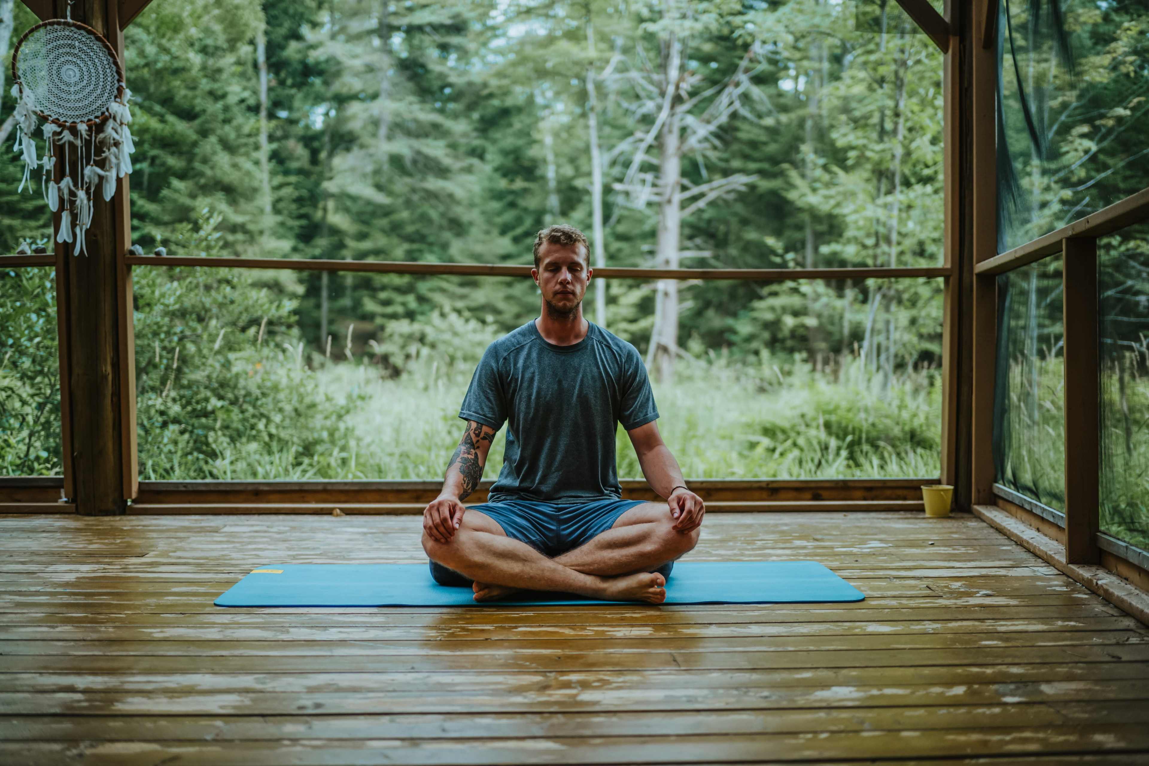A man sits cross-legged on a yoga mat in a wooden cabin surrounded by trees.