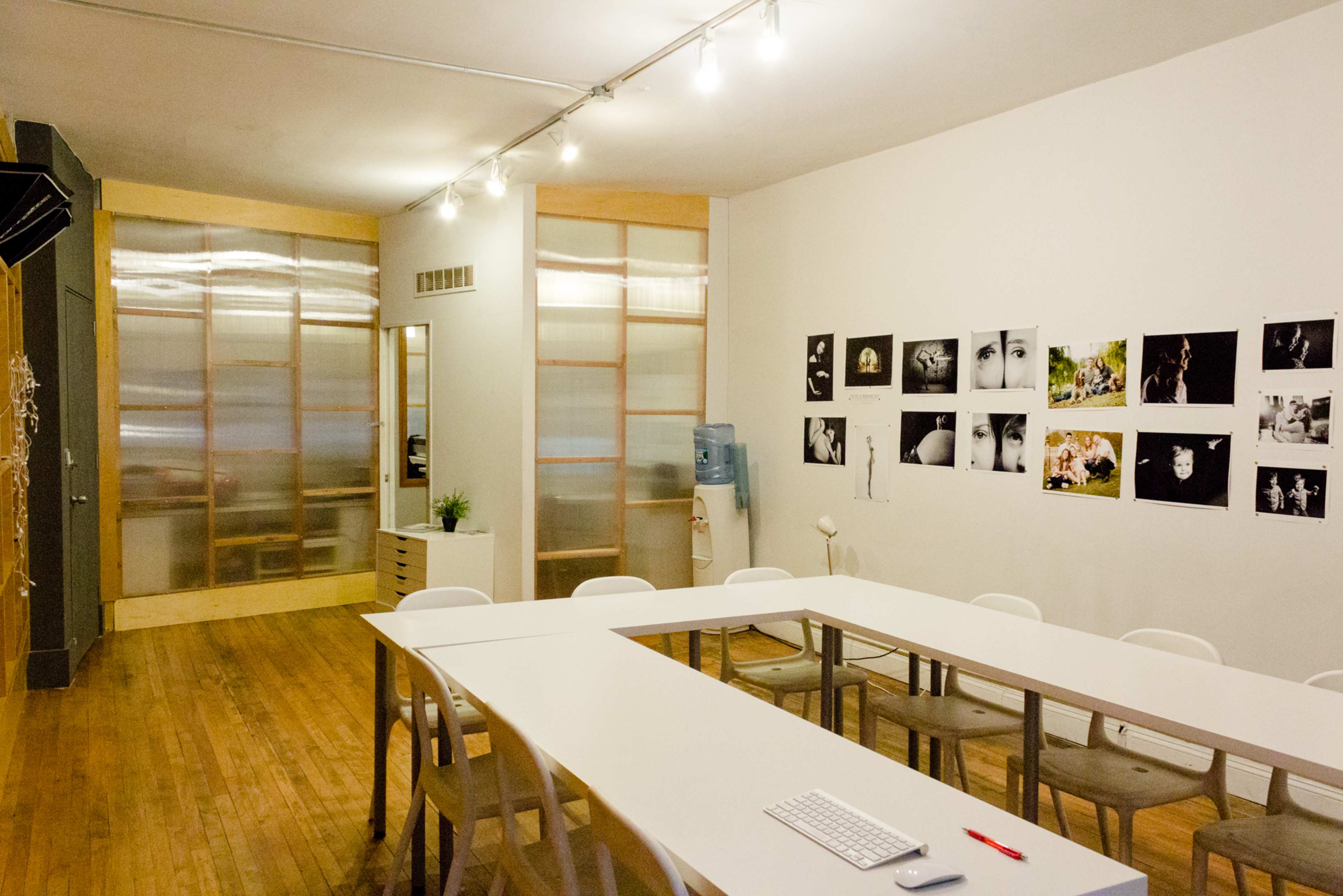 A well-lit room features a large white table surrounded by chairs, with a water cooler and a series of framed photographs on the wall.