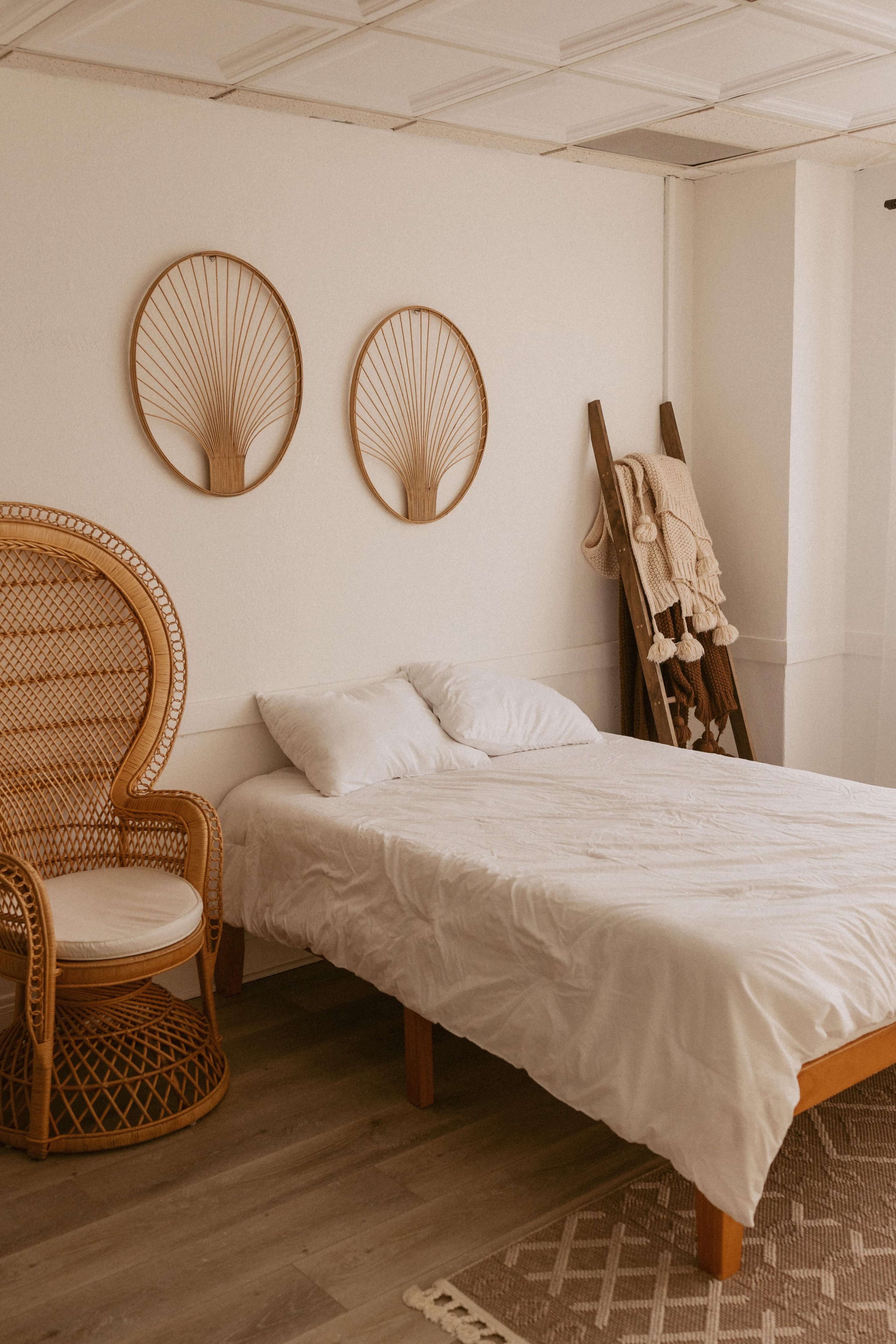 The image shows a minimalist bedroom featuring a bed with white bedding, a rattan chair, and decorative wall art made of woven materials.