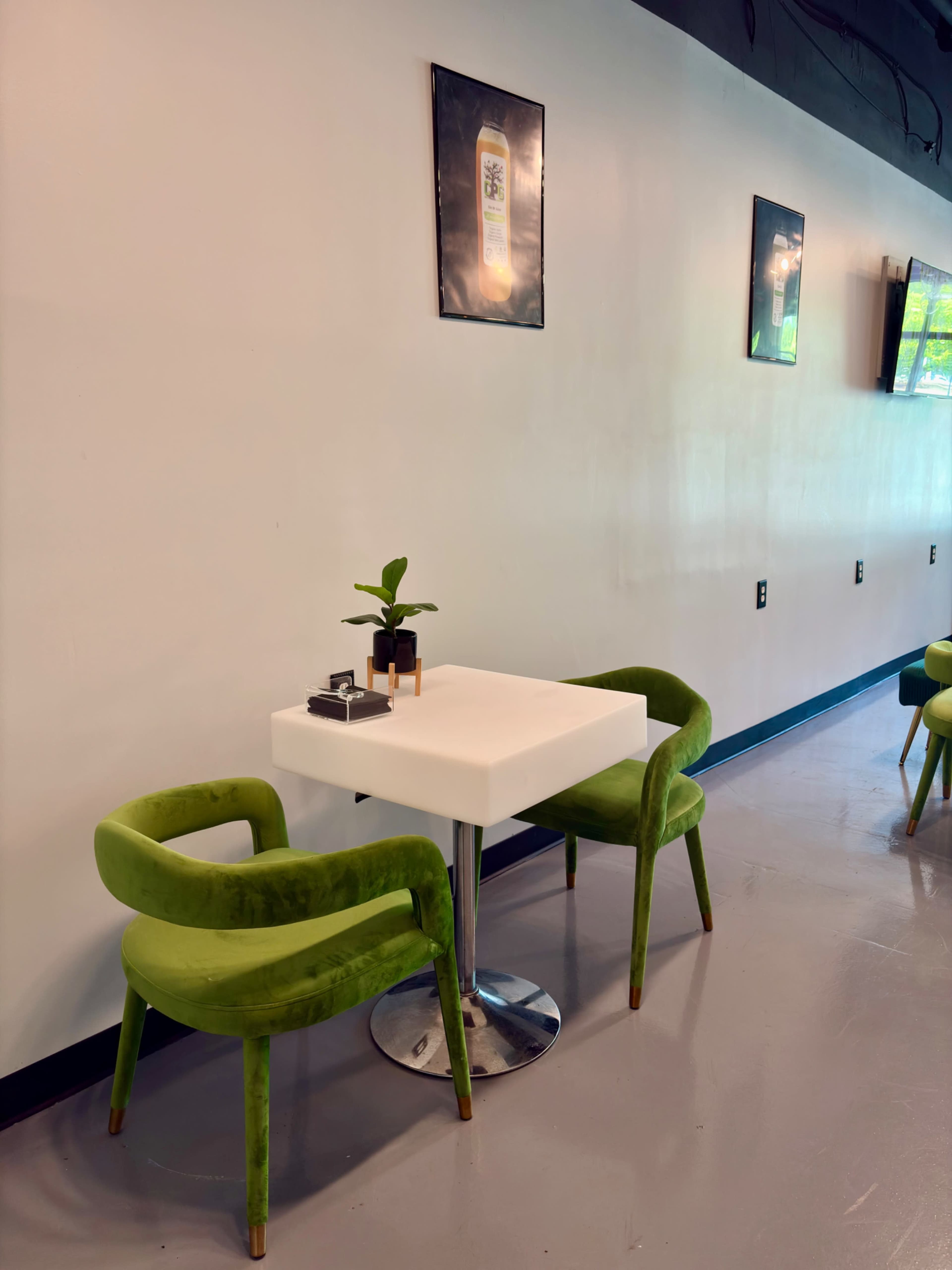 A small white table with two green chairs and a potted plant sits against a plain white wall, with framed pictures hanging above.