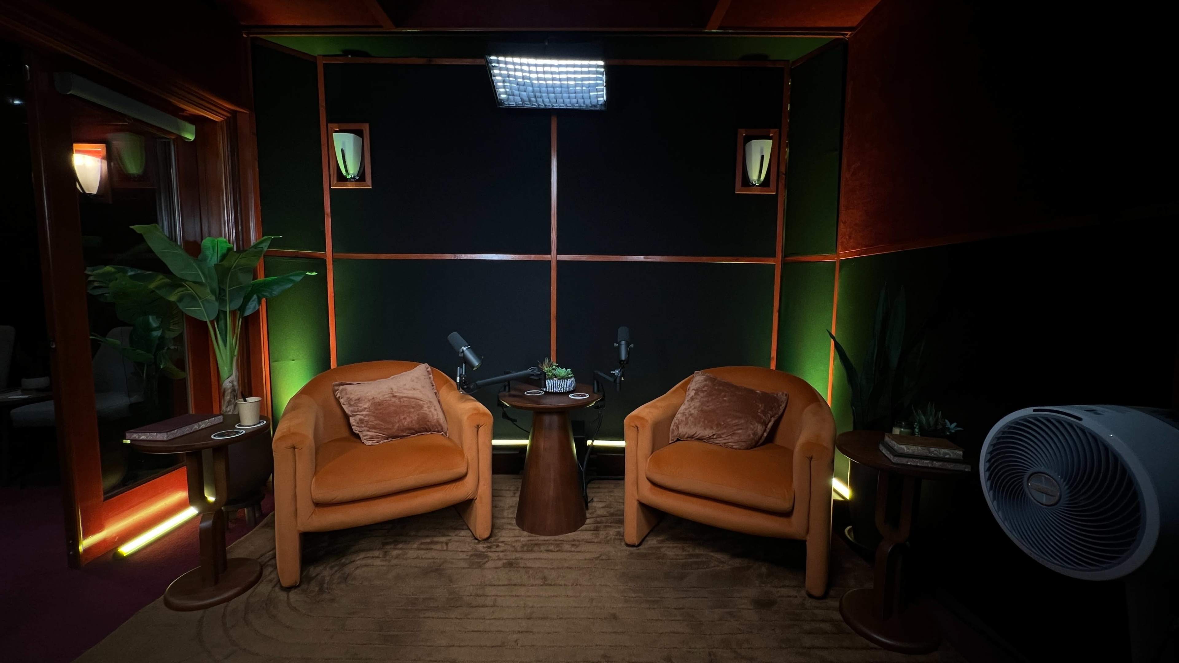The image shows a cozy recording studio setup with two orange chairs facing a small round table, surrounded by greenery and softly lit walls.