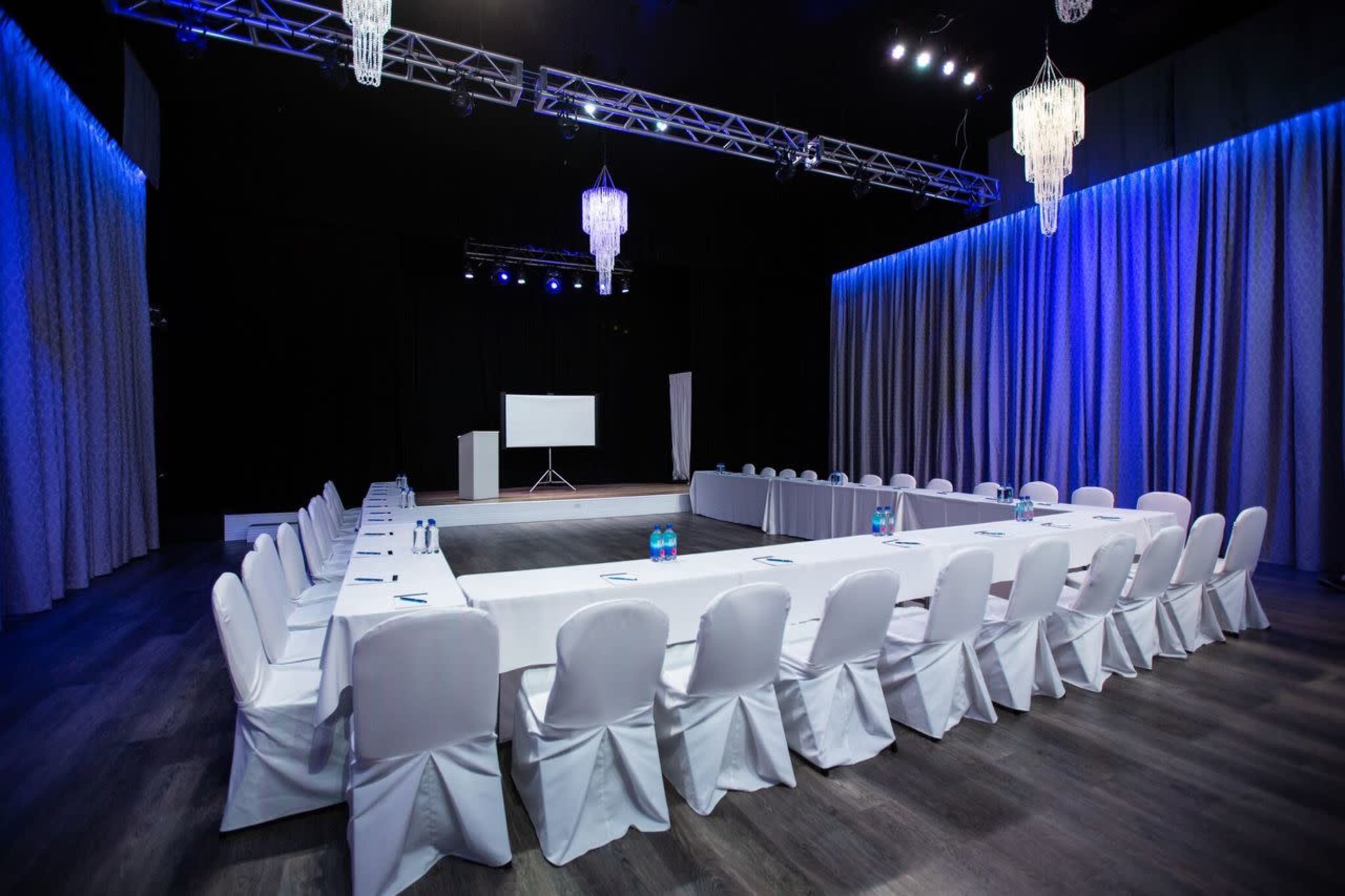 The image shows a conference room arranged with a U-shaped seating layout, featuring white tablecloths and chairs, under blue lighting and hanging chandeliers.