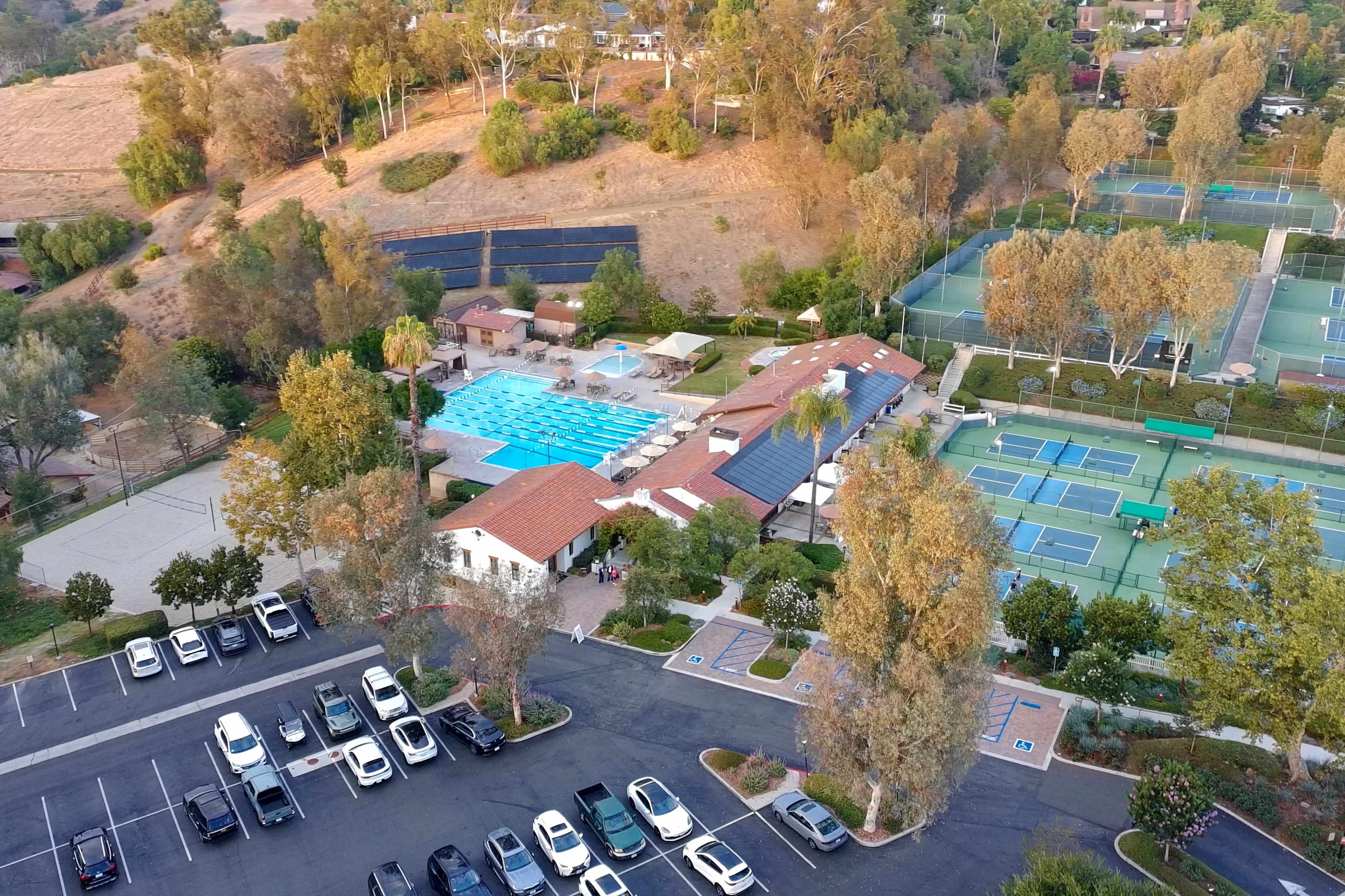 An aerial view shows a multi-facility complex with a swimming pool, tennis courts, and a parking area filled with cars, surrounded by trees and hills.