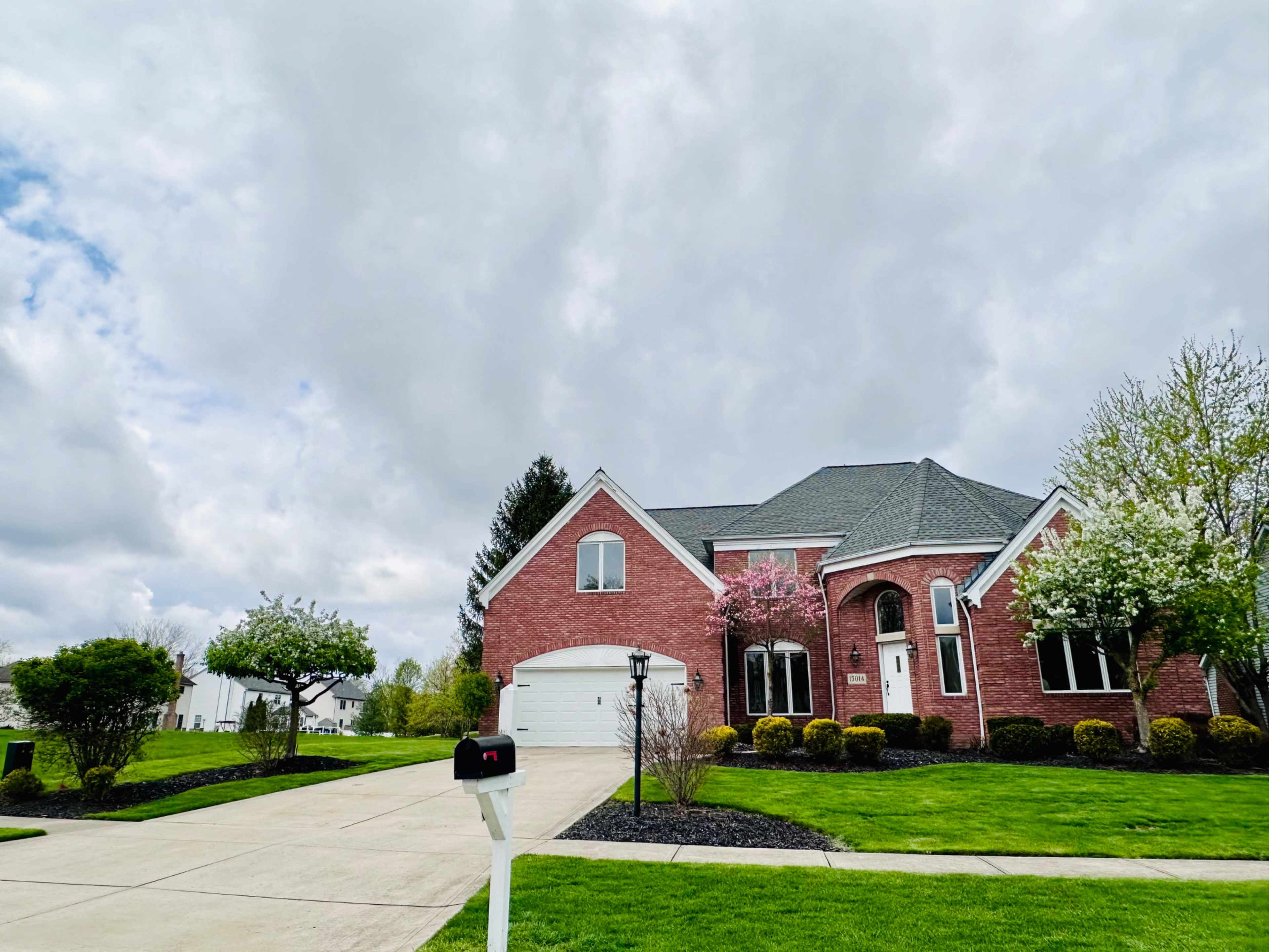 A red-brick two-story house with a large front yard and driveway is set against a cloudy sky.