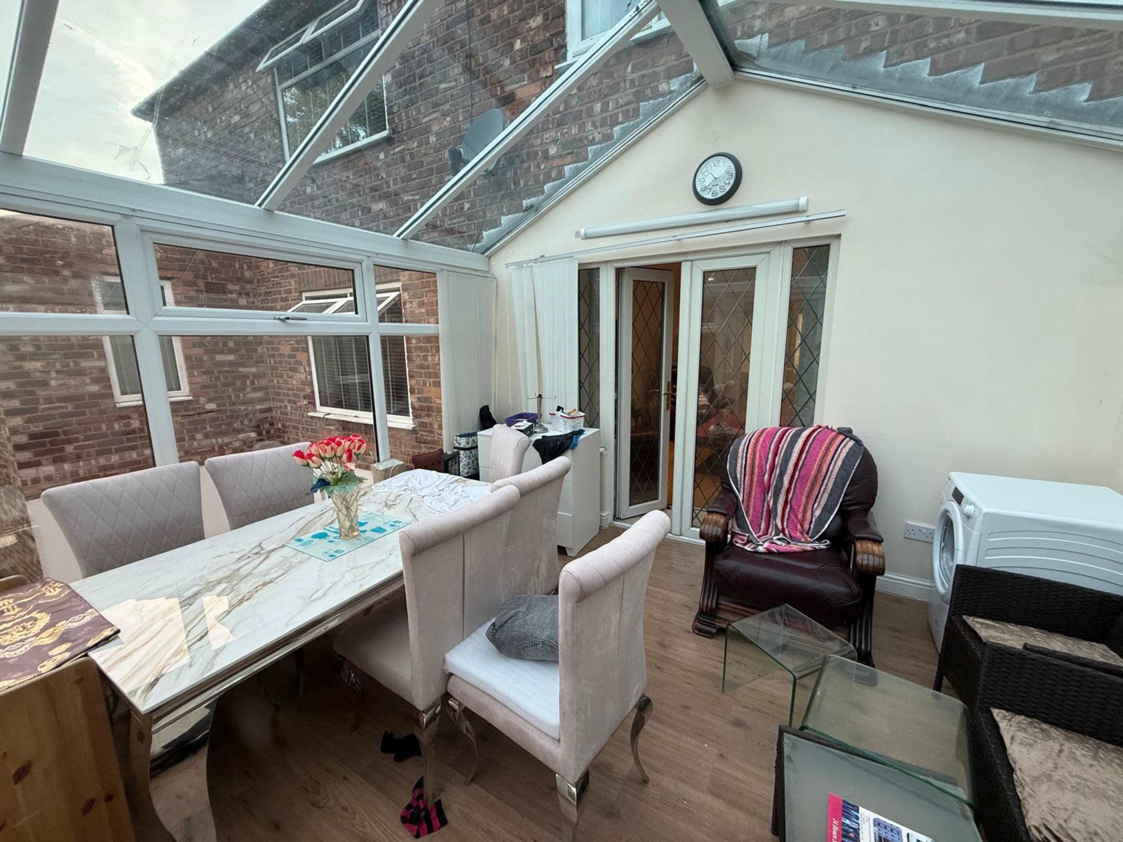 The image shows a bright conservatory with a dining table, pink upholstered chairs, a recliner, and a washer in the corner.