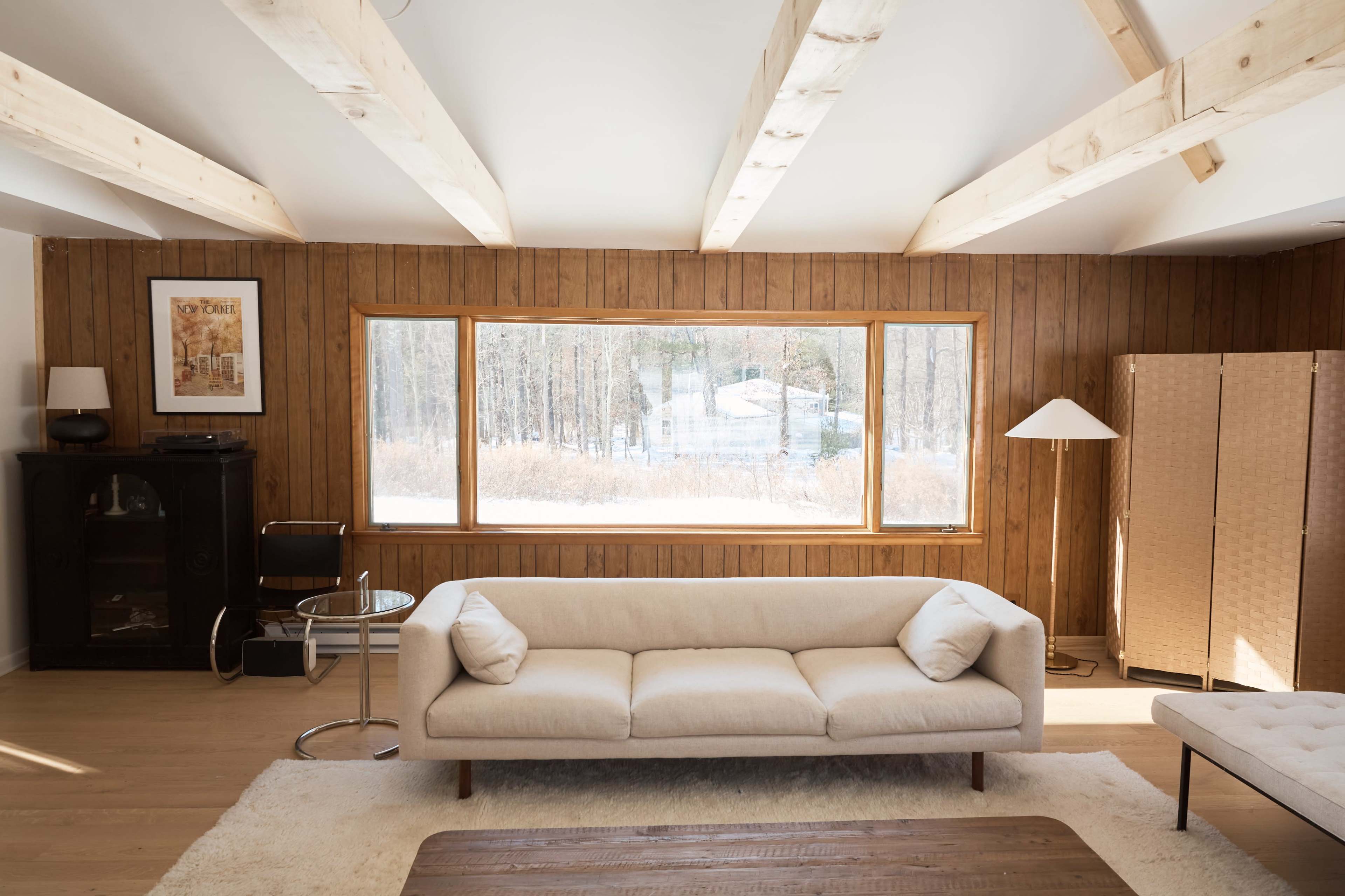 A light-filled living room features a beige sofa, a wooden table, and large windows overlooking a snowy landscape.