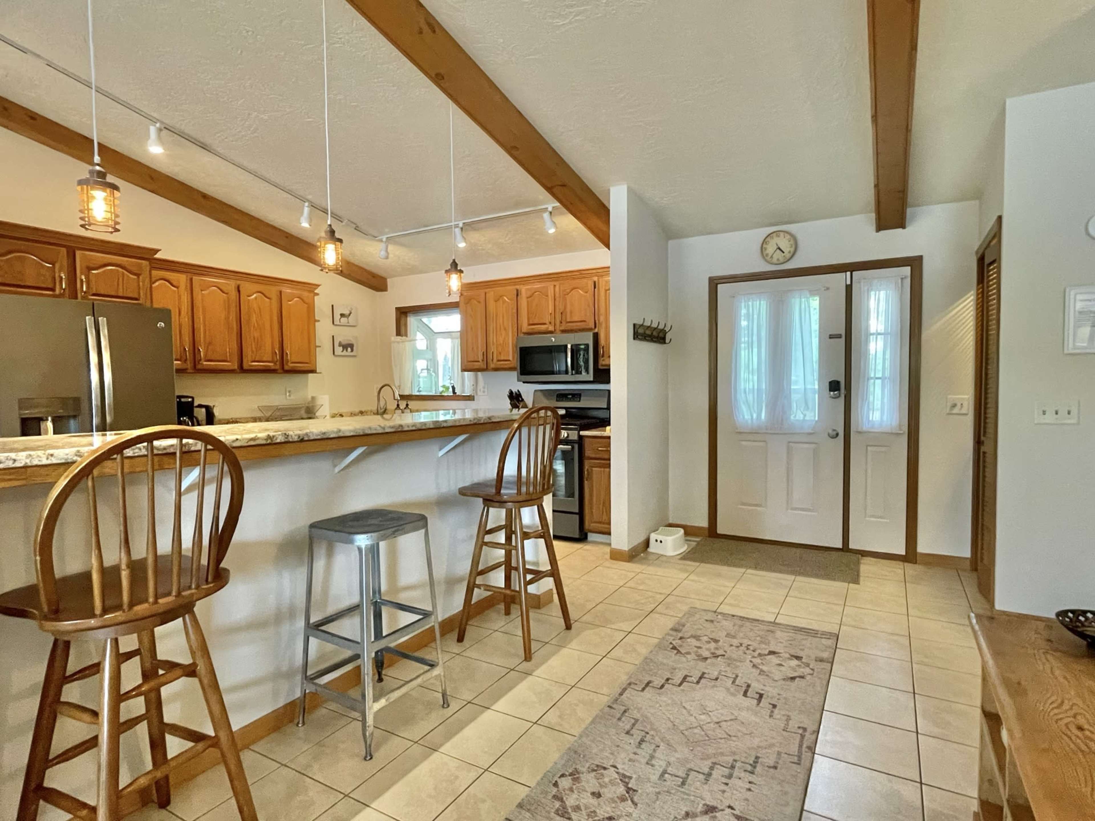 A spacious kitchen with wooden cabinets, a granite countertop, and two barstools, adjacent to an entryway featuring a front door and a small rug.