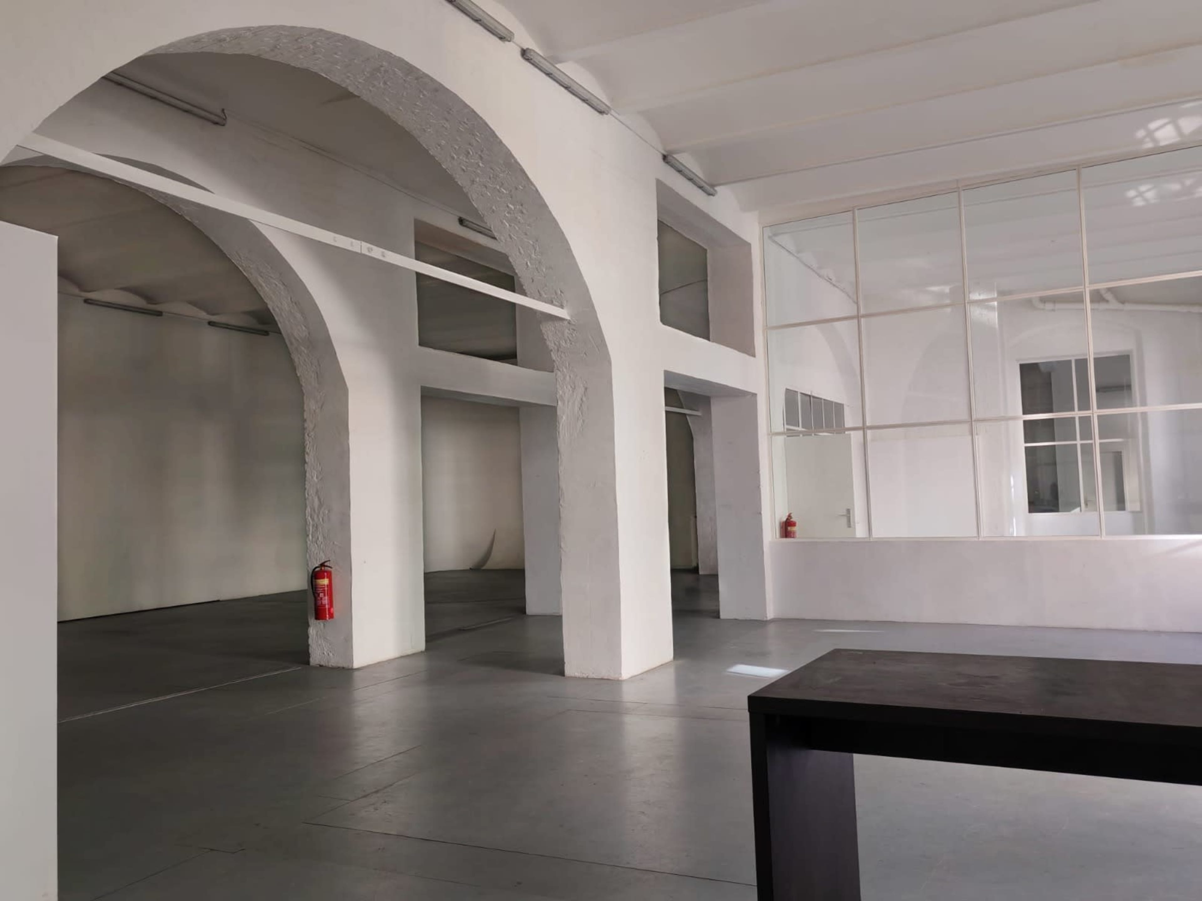 The image shows a spacious interior with white walls, high ceilings, and large arched openings, featuring a few fire extinguishers and minimal furniture.