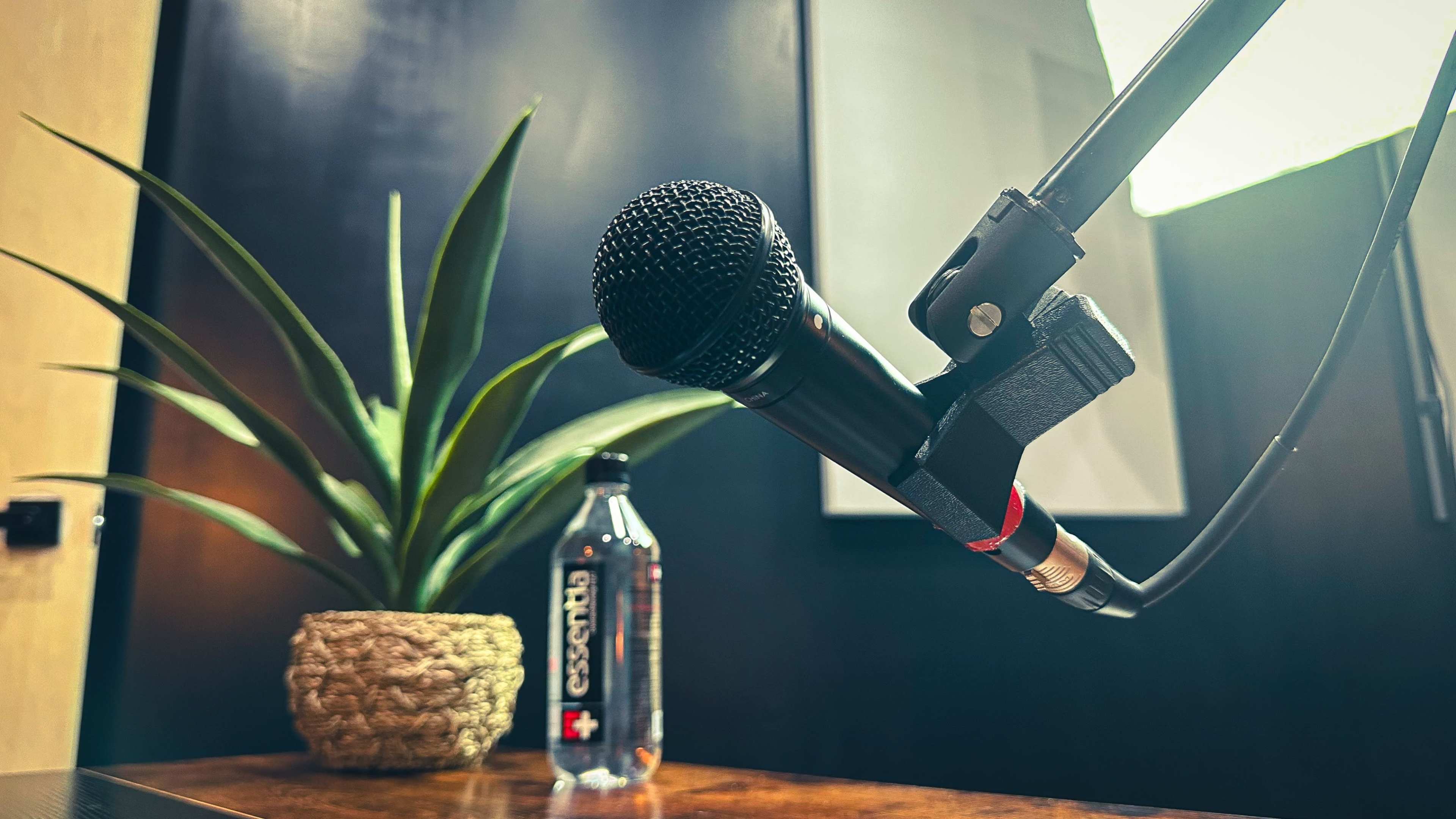 A microphone is positioned in front of a potted plant and a bottle of water on a wooden table.