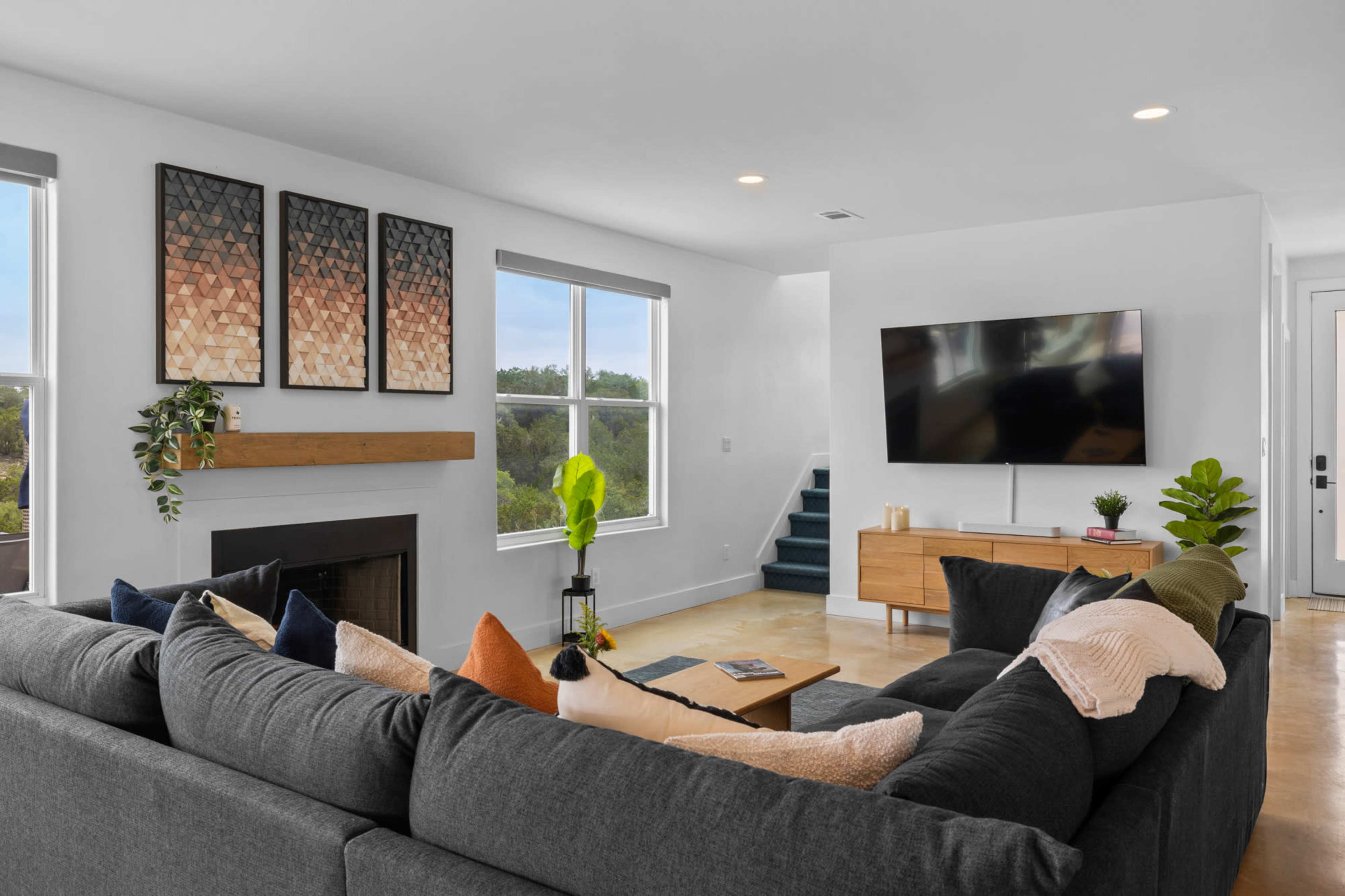 A modern living room features a large gray sectional sofa, a wooden coffee table, a flat-screen TV mounted on the wall, and large windows providing natural light.