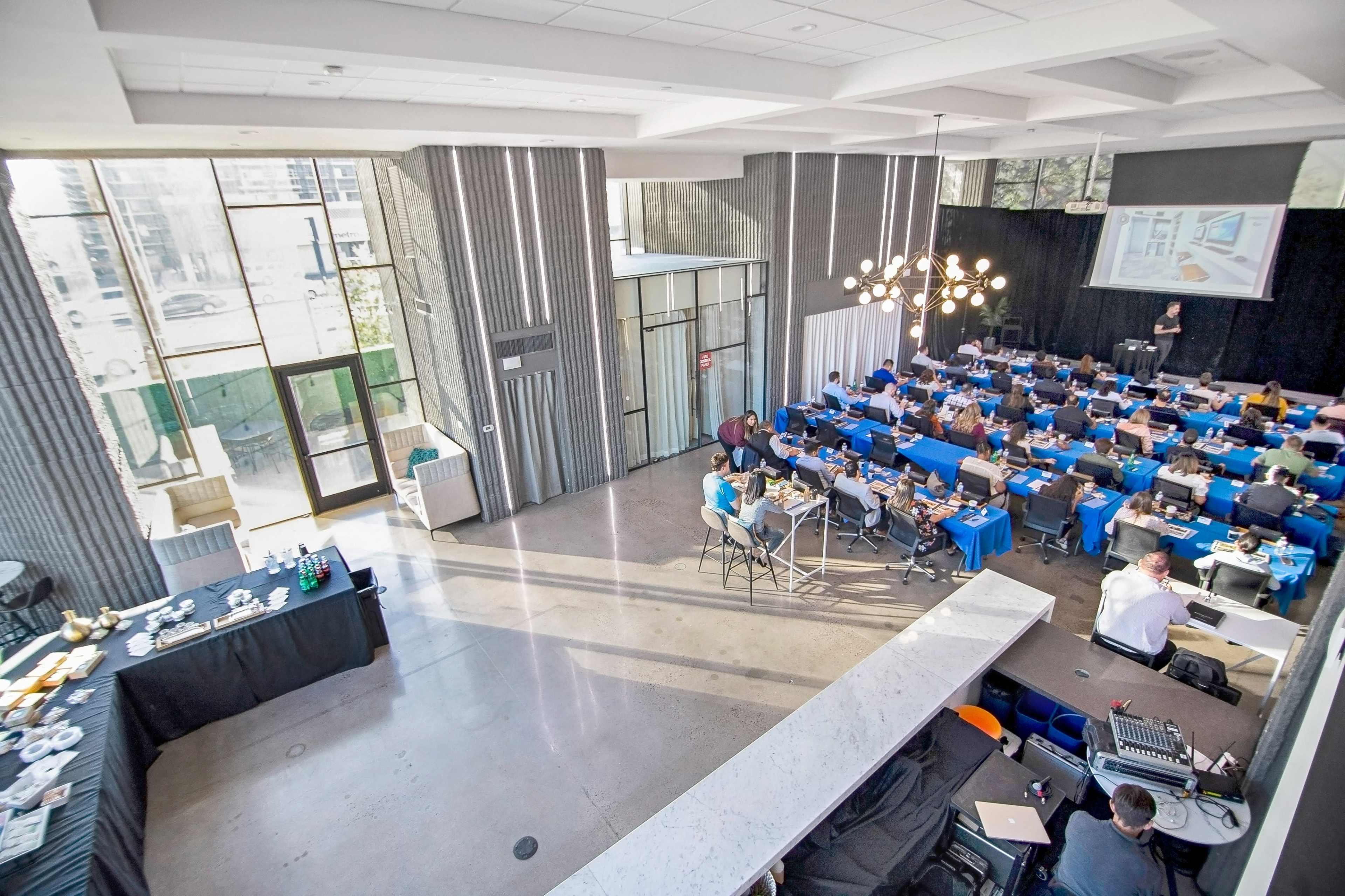 A well-attended conference room with rows of tables set up for participants, including equipment and refreshments, while a speaker presents at the front.
