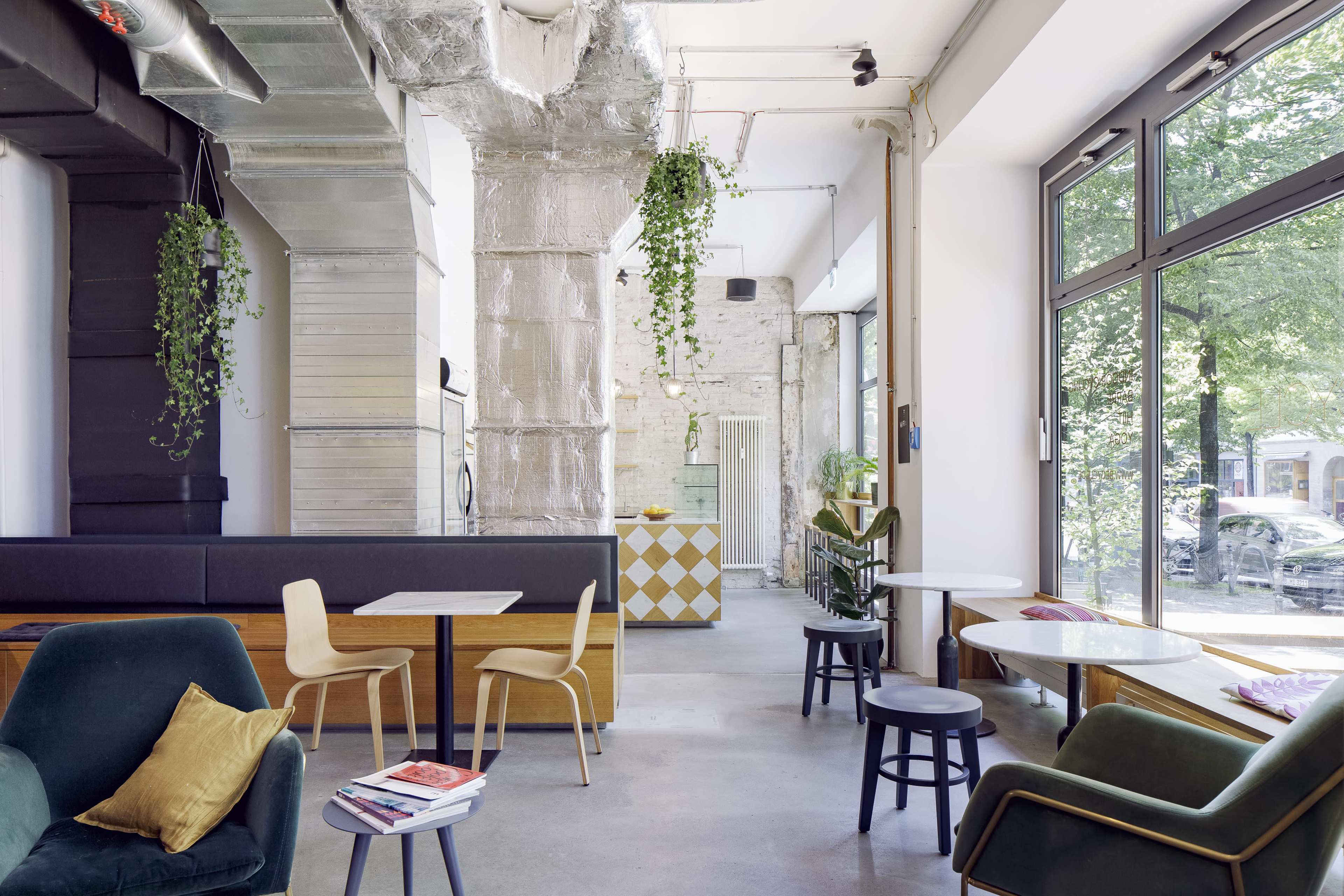 A modern café interior with large windows, a mix of seating options, and greenery hanging from the ceiling.