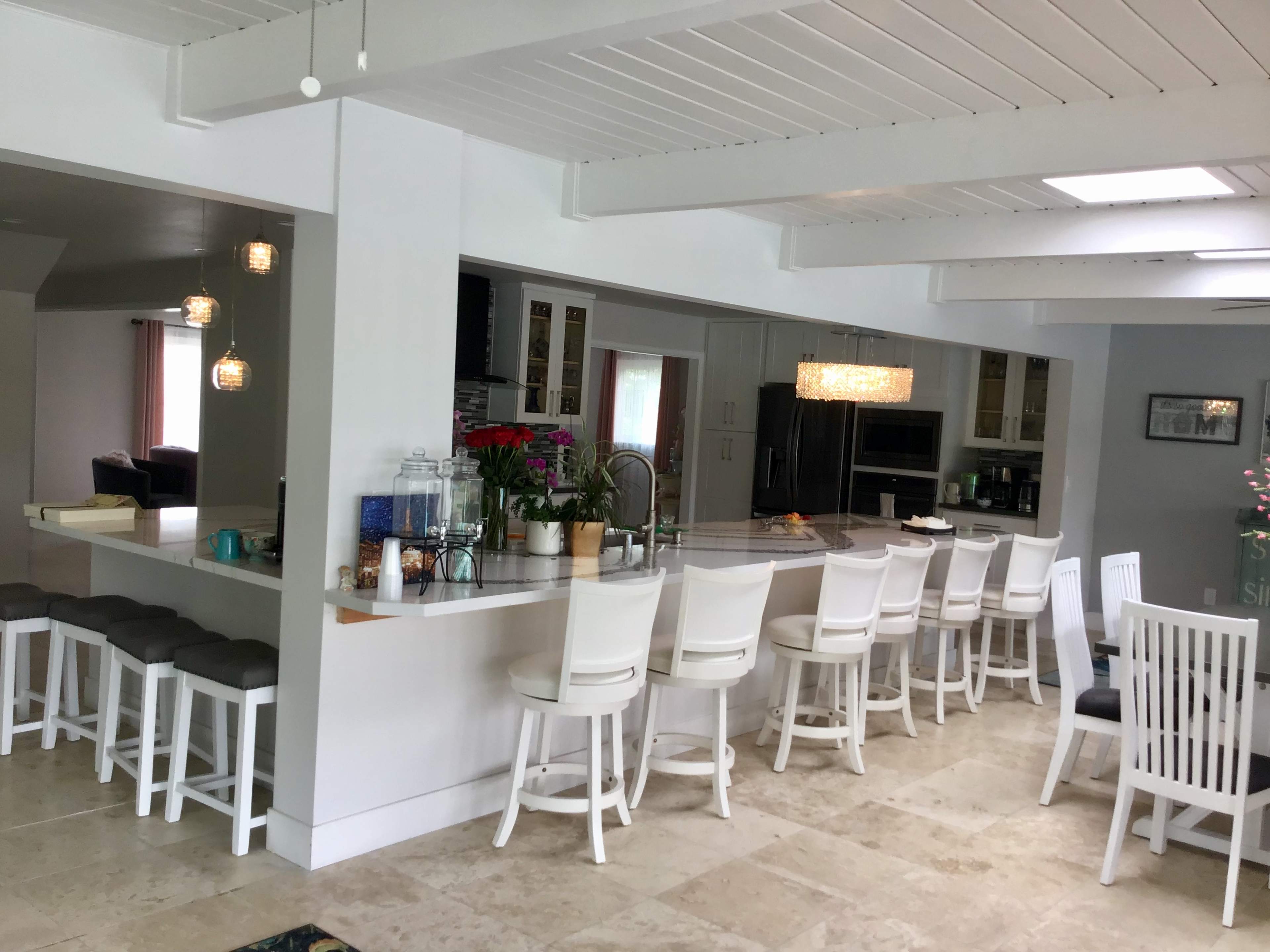 The image shows a modern kitchen with a long, white countertop and several white barstools arranged along one side.
