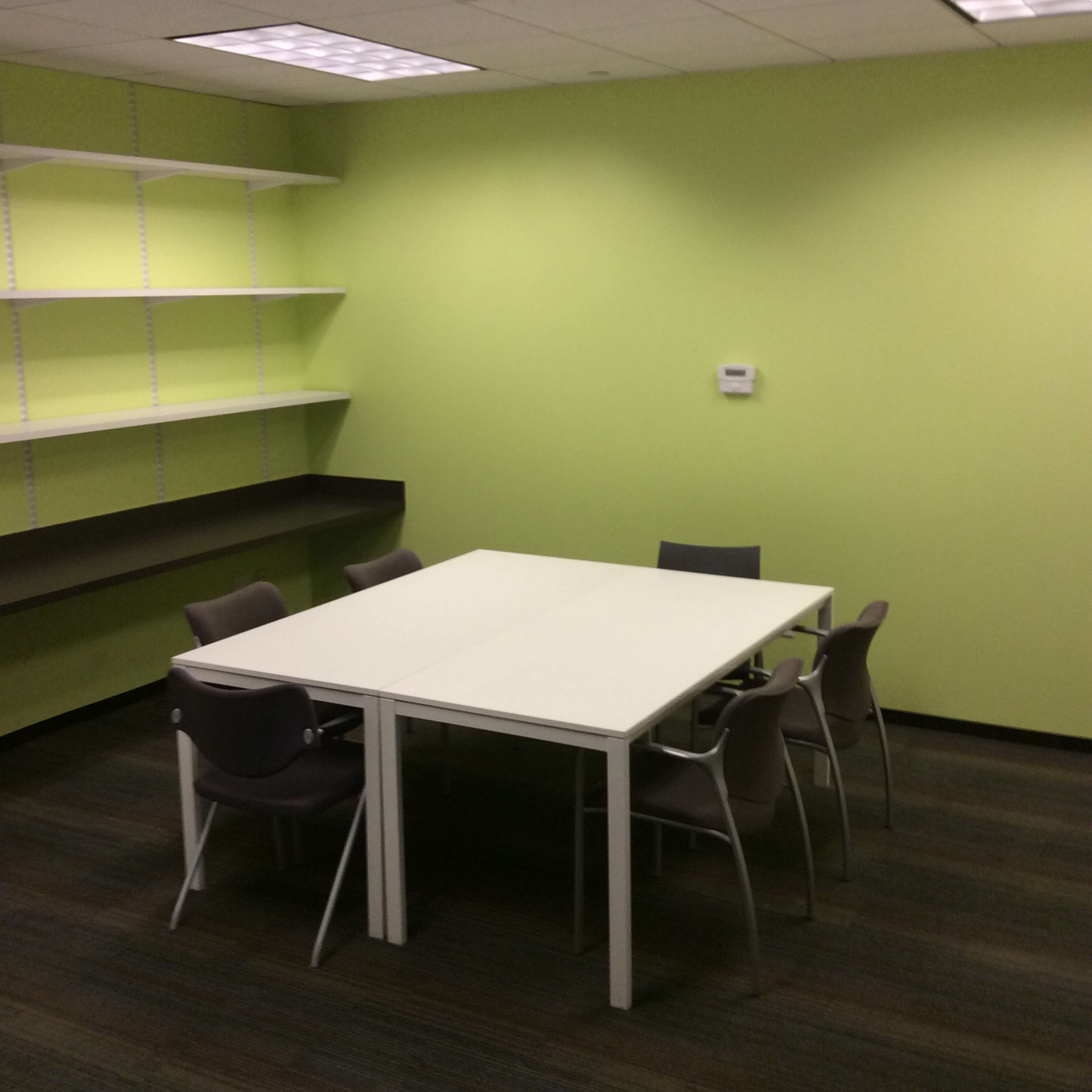 A small conference room with a white table and six chairs, set against a bright green wall and a shelf on one side.