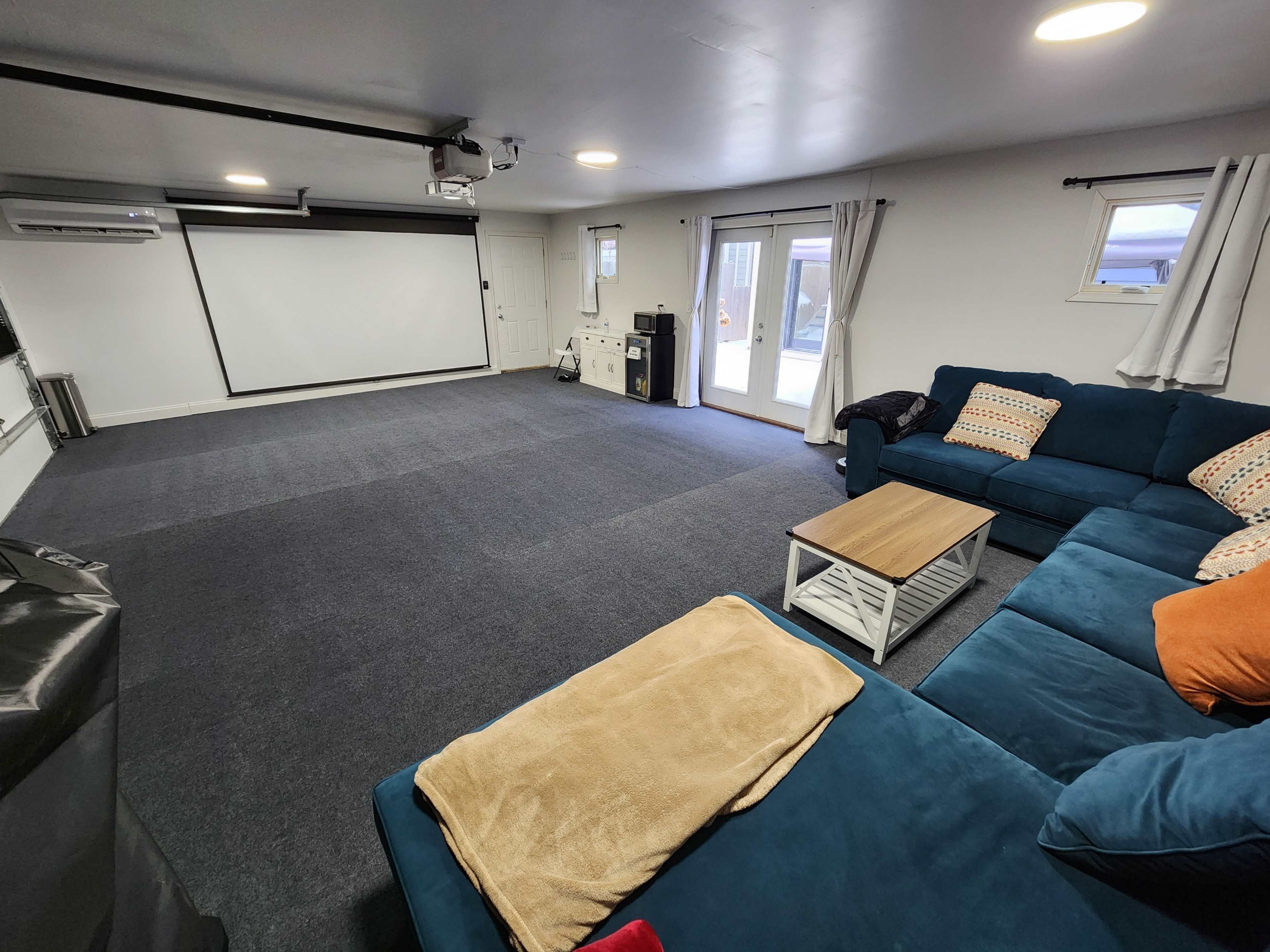 A spacious room features a blue sectional sofa, a coffee table, and a large white projection screen against one wall.