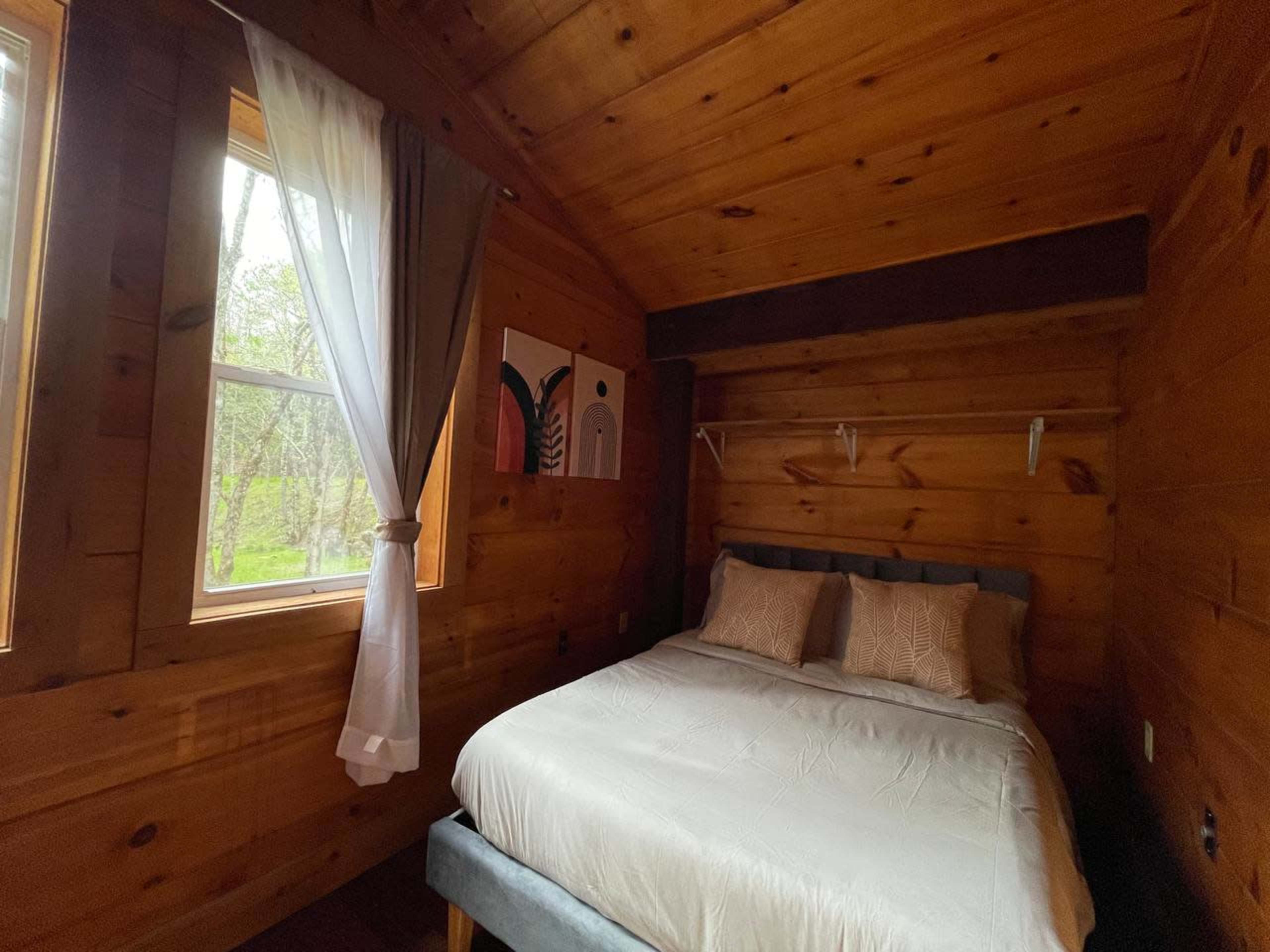The image shows a small bedroom with wooden walls, a bed with light bedding, and windows allowing sunlight to enter.