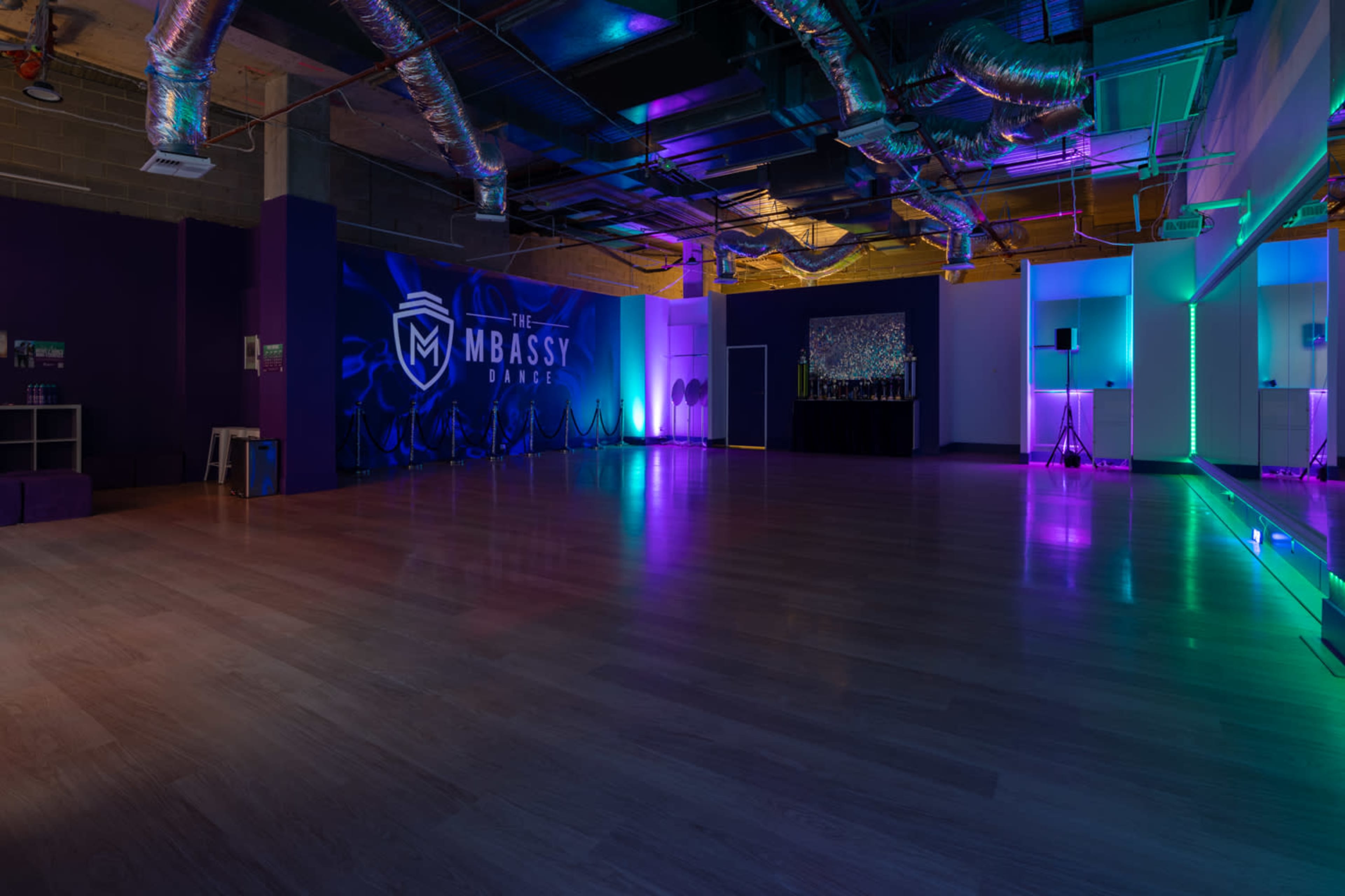 A spacious dance studio with a polished wooden floor, illuminated by colorful lighting in purple and blue tones, and featuring a branded wall and a mirrored area.