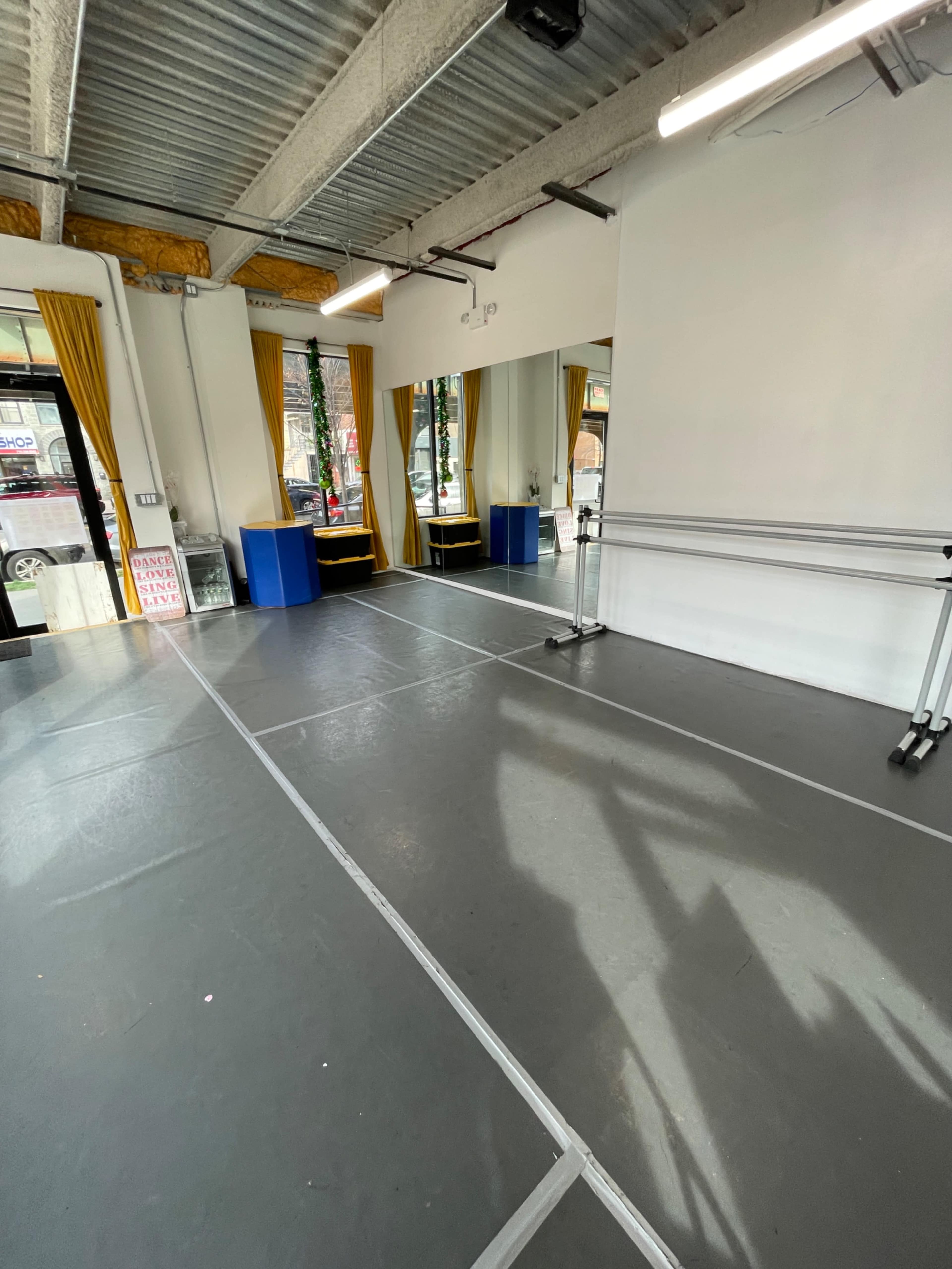 An empty dance studio with gray flooring, large windows draped with yellow curtains, and mirrored walls.