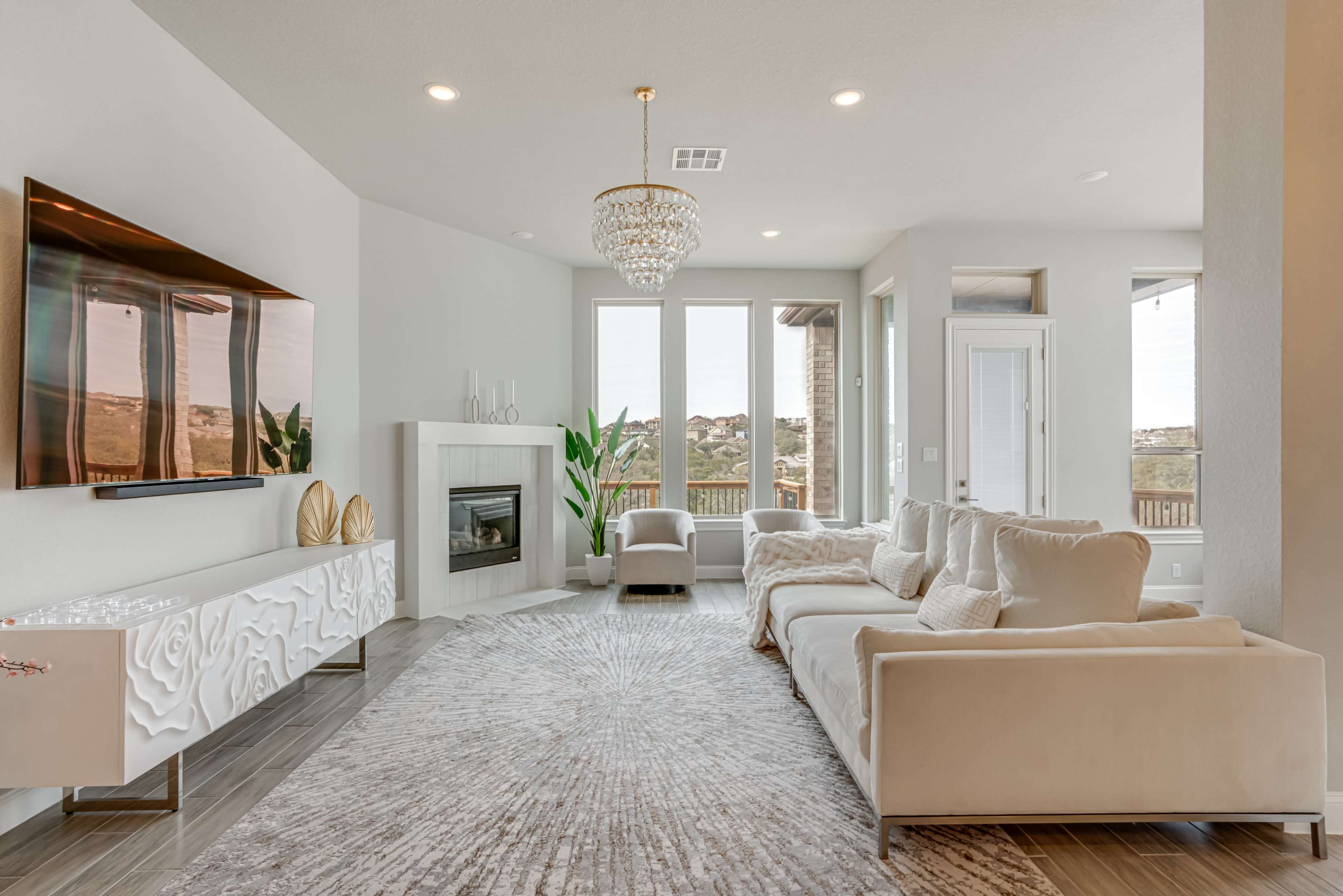 The living room features a modern design with a large sectional sofa, a flat-screen TV mounted on the wall, a fireplace, and large windows overlooking a scenic view.