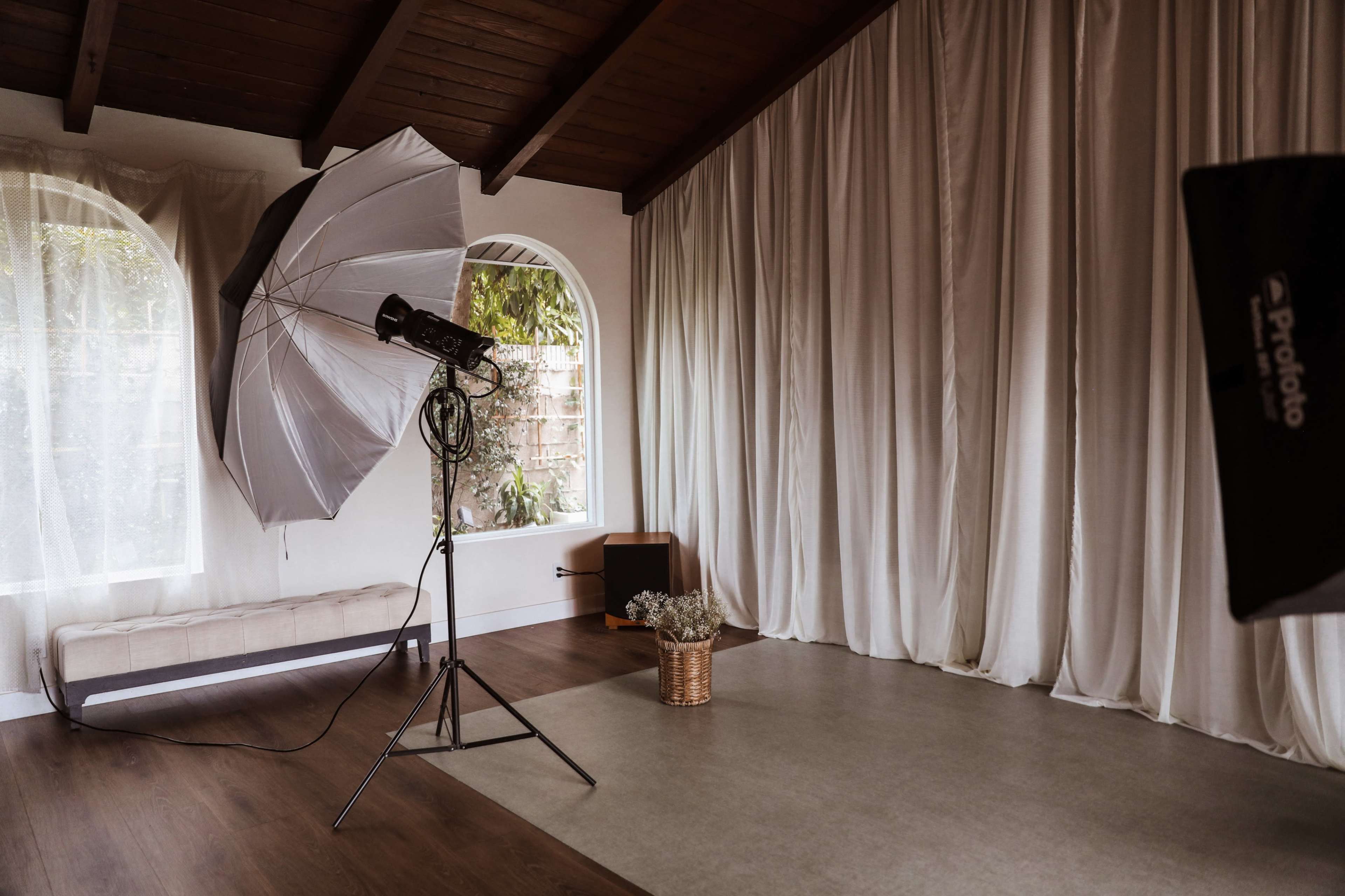 The image shows a photography studio with a large umbrella softbox, a woven basket of flowers, and draped white curtains against a wooden ceiling.