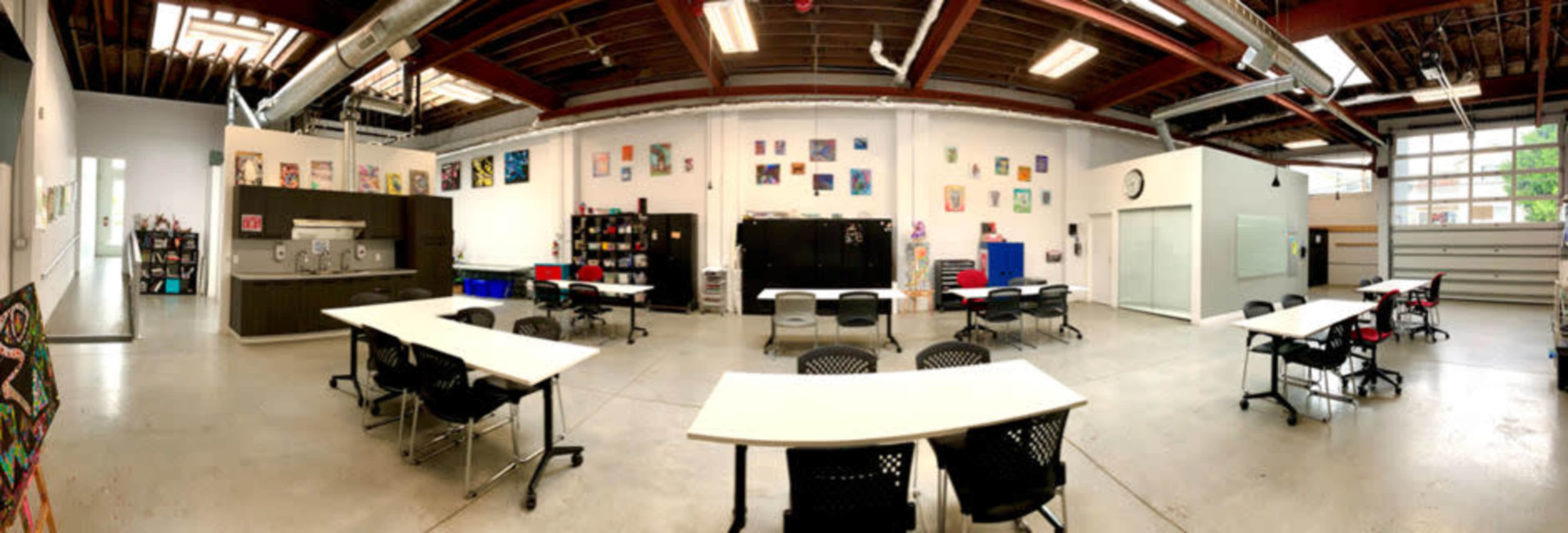 A spacious, well-lit classroom with several tables and chairs arranged in a U-shape, along with various artworks displayed on the walls.