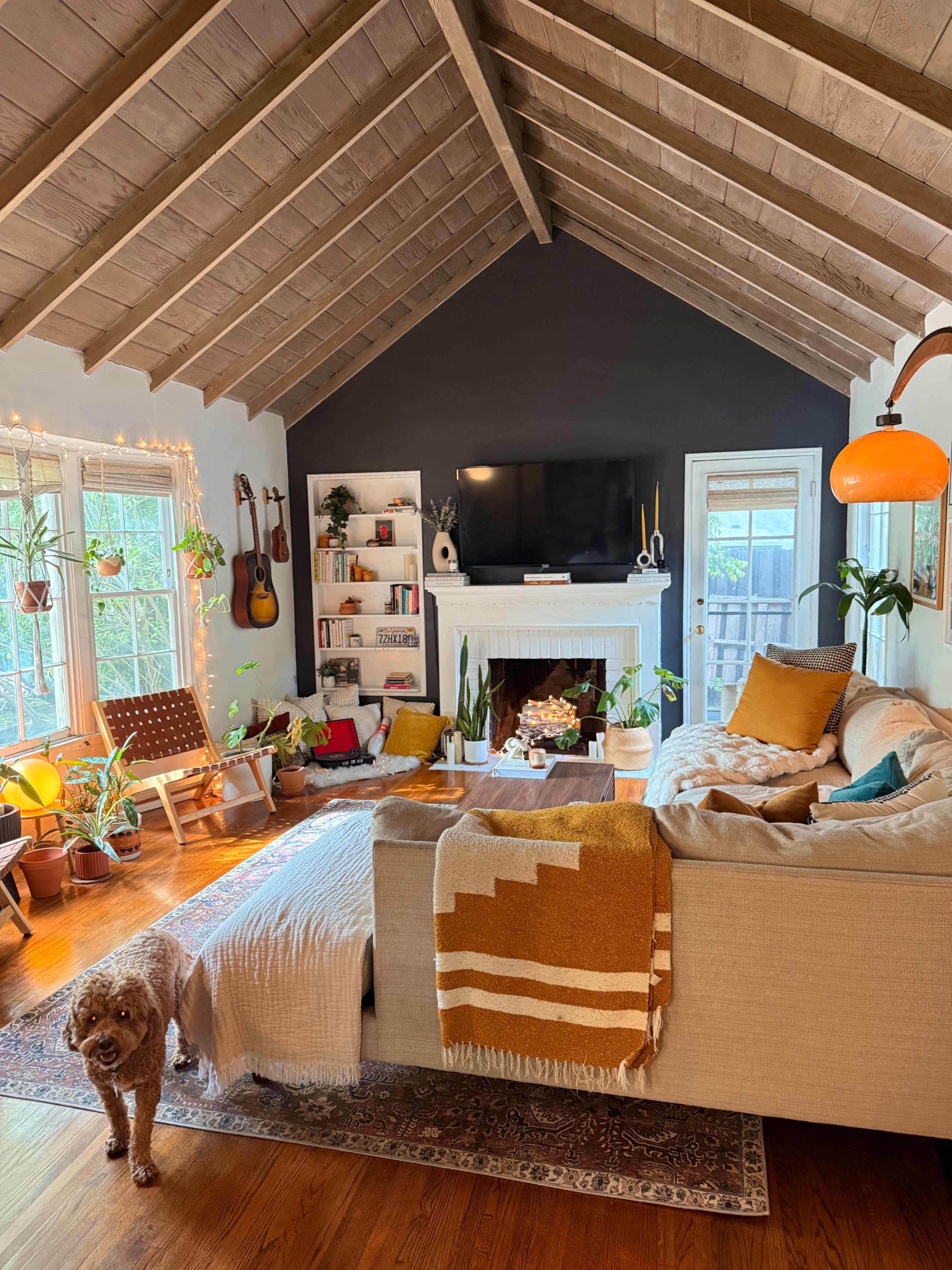 The living room features a high, slanted ceiling with wooden beams, a large sofa adorned with blankets, plants throughout, and a cozy seating area with a guitar and a dog nearby.