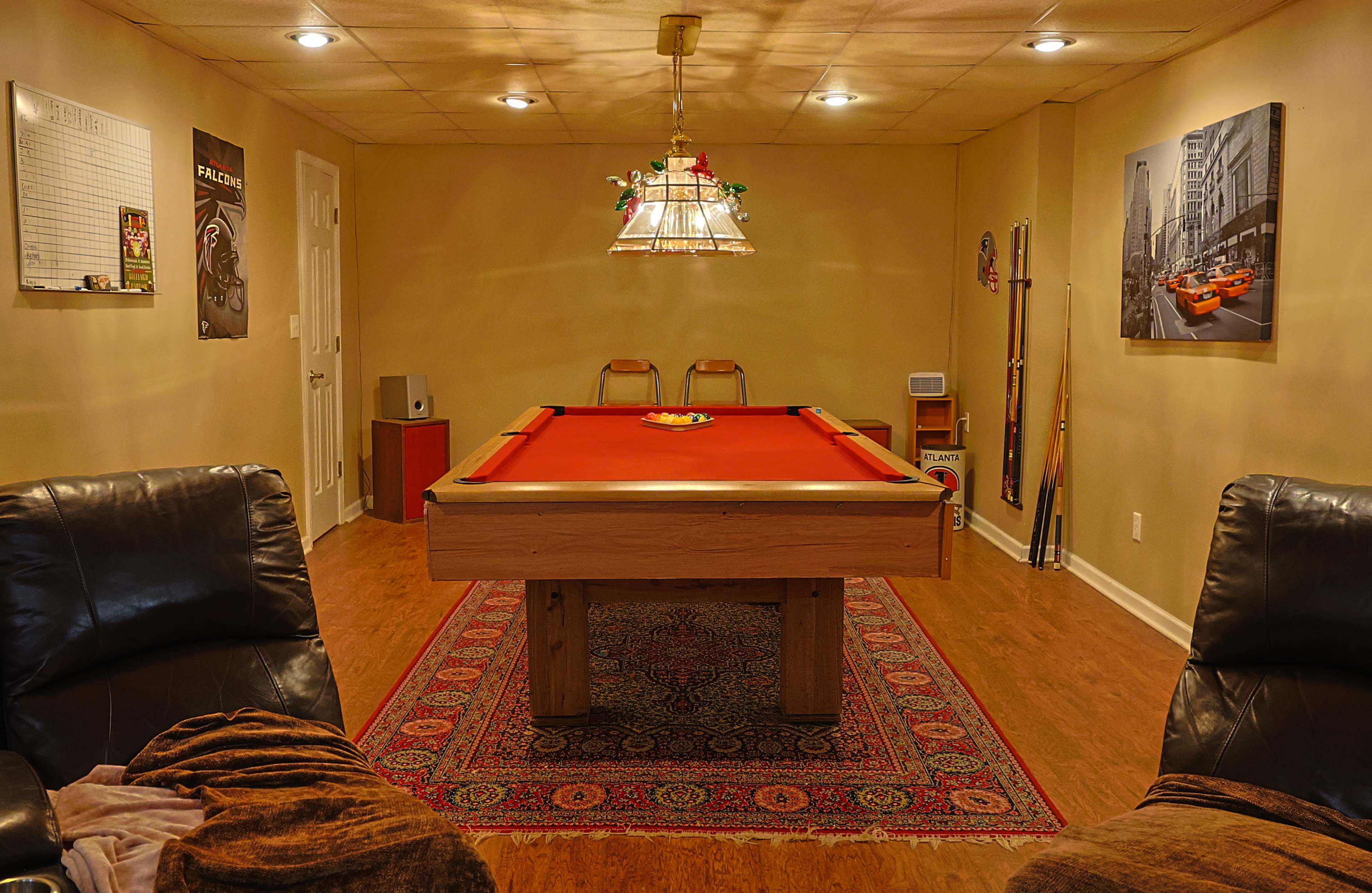 The image shows a pool table in a well-lit basement room, flanked by two recliners and decorated with a rug and wall art.