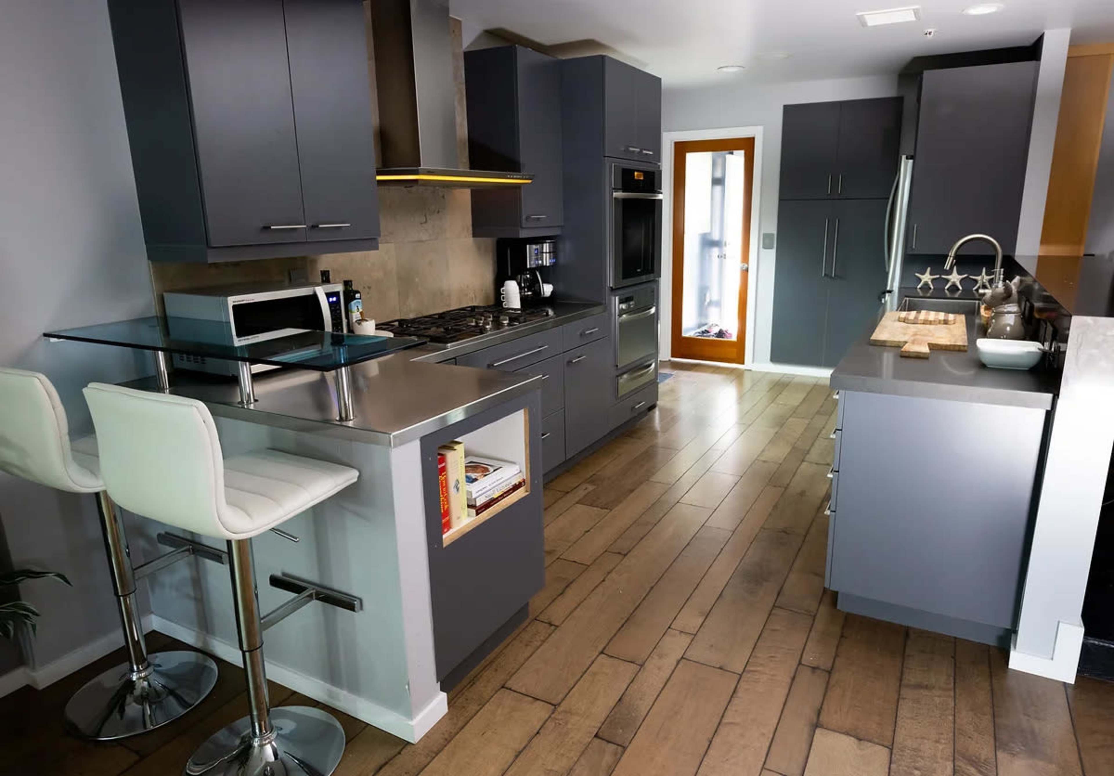 A modern kitchen with gray cabinets, stainless steel appliances, a bar with two white stools, and a wooden floor.